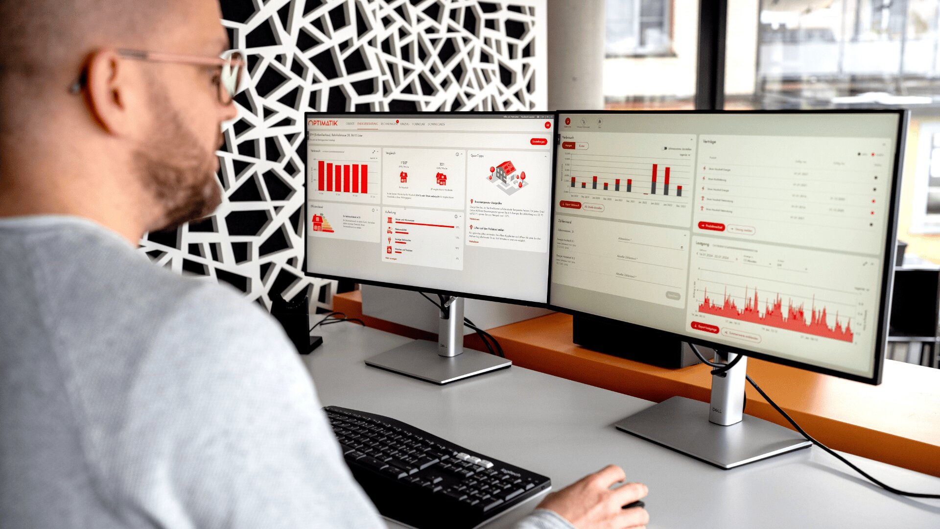 Eine Person sitzt an einem Schreibtisch mit Tastatur und Maus und betrachtet Datentabellen und Diagramme auf zwei großen Computermonitoren in einer modernen Büroumgebung.