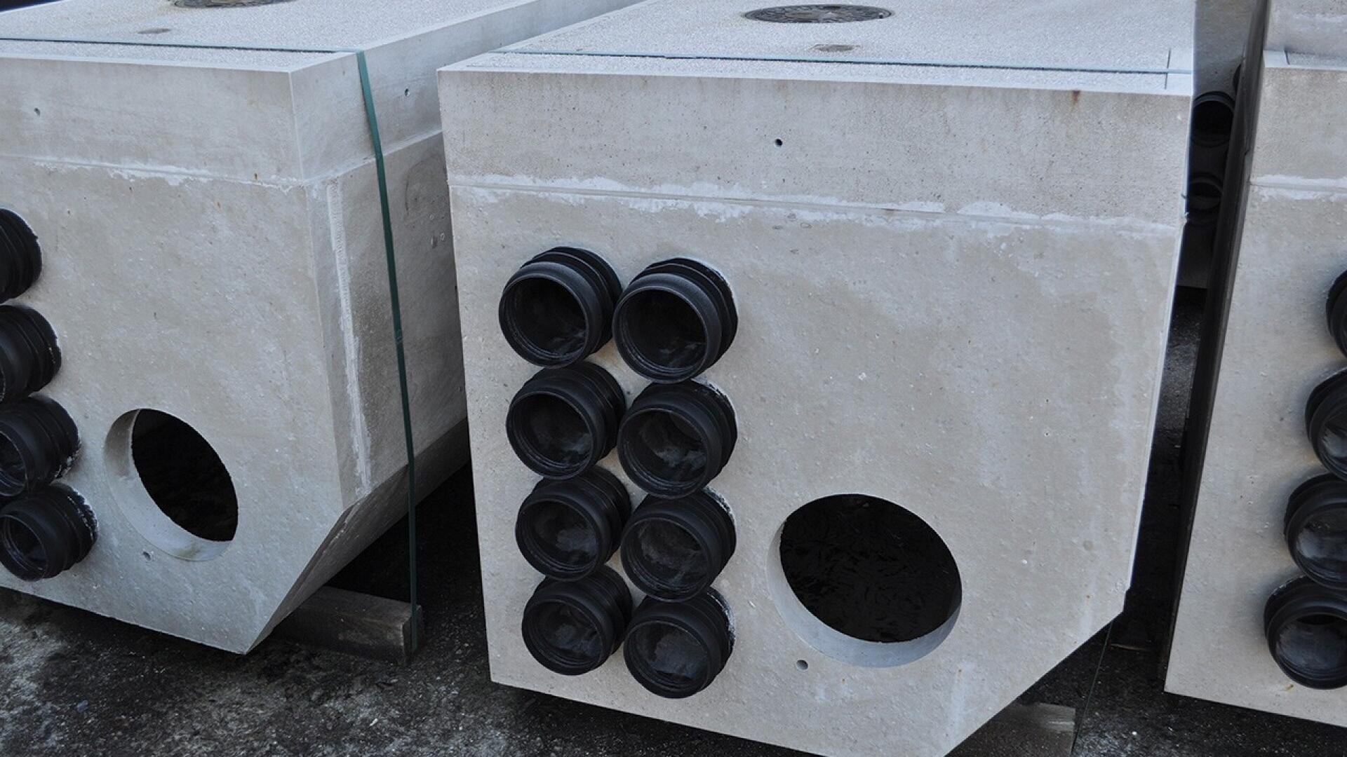 Large concrete blocks with multiple round black pipe fittings and a large circular opening stacked outdoors on a concrete surface.