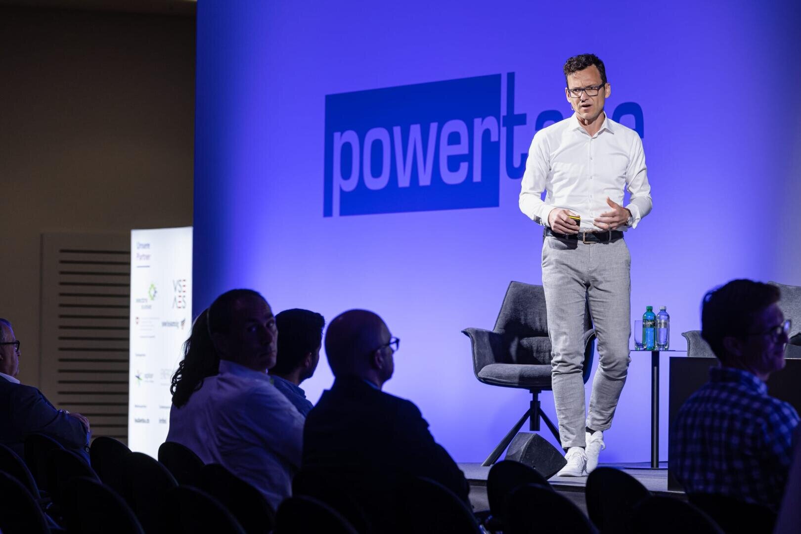 Ein Mann in einem weißen Hemd und einer grauen Hose steht auf der Bühne und hält Papiere in der Hand, hinter ihm ein blaues Powertalk-Schild. Mehrere Personen sitzen im Publikum, und auf der Bühne stehen zwei Stühle und Wasserflaschen.