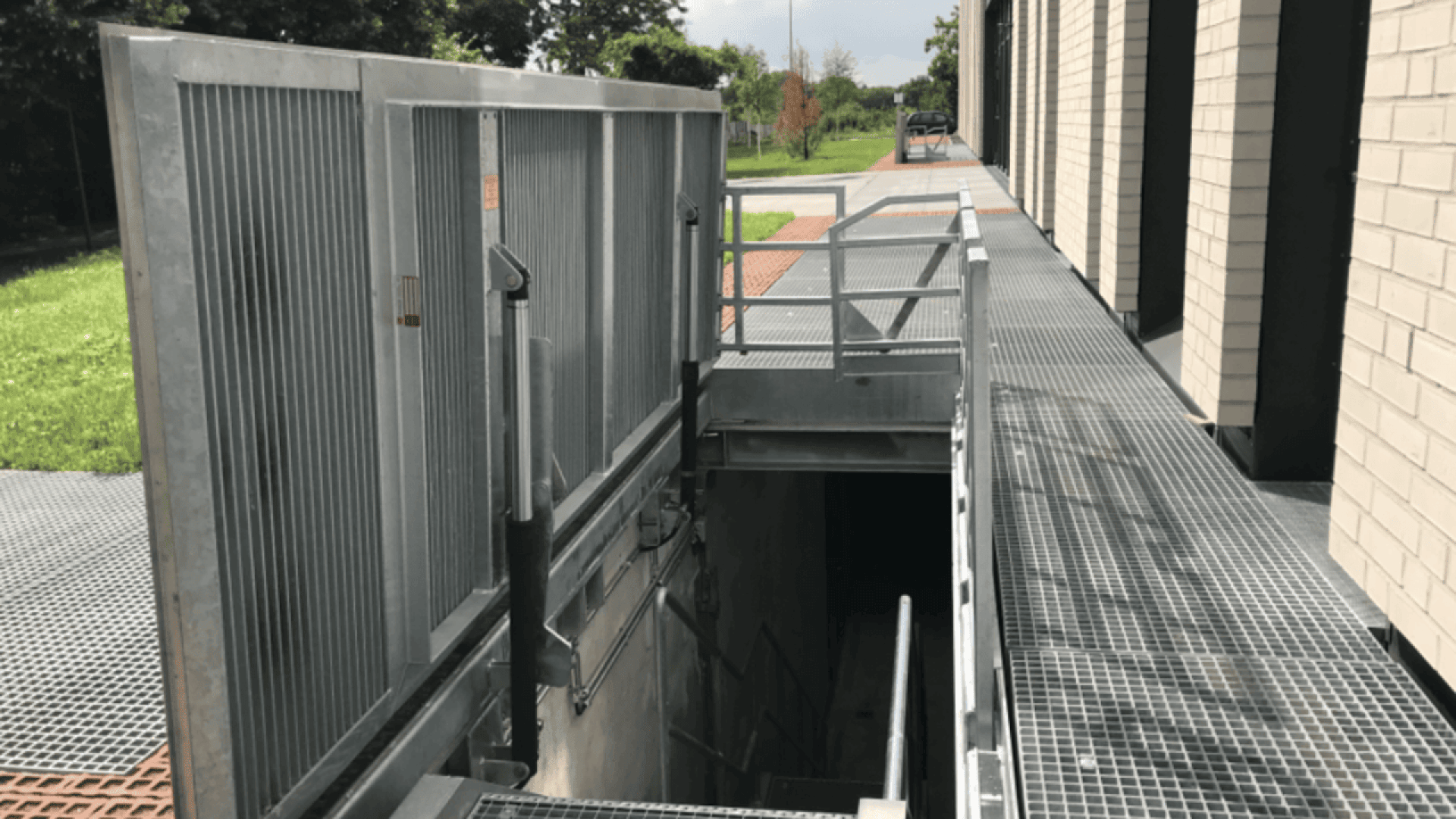 A metal flood barrier gate partially opens to reveal stairs leading down to a lower level, next to a building with a grated walkway and green grass in the background.