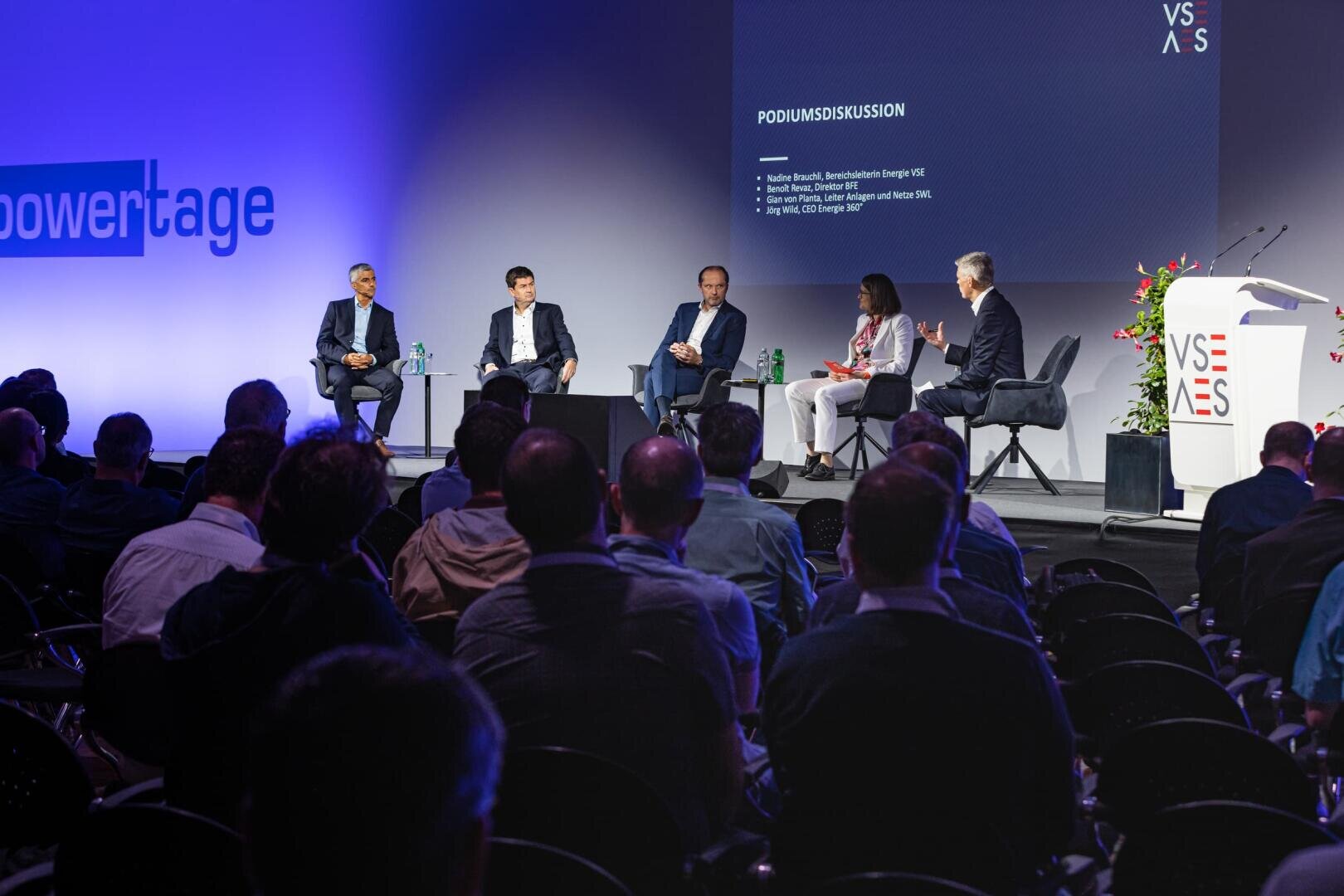 Fünf Personen sitzen bei einer Podiumsdiskussion auf einer Konferenz auf der Bühne und blicken auf das Publikum. Auf einem großen Bildschirm werden die Details der Diskussion angezeigt, und die Bühne ist gut beleuchtet, mit einem Podium und Blumen auf der rechten Seite.