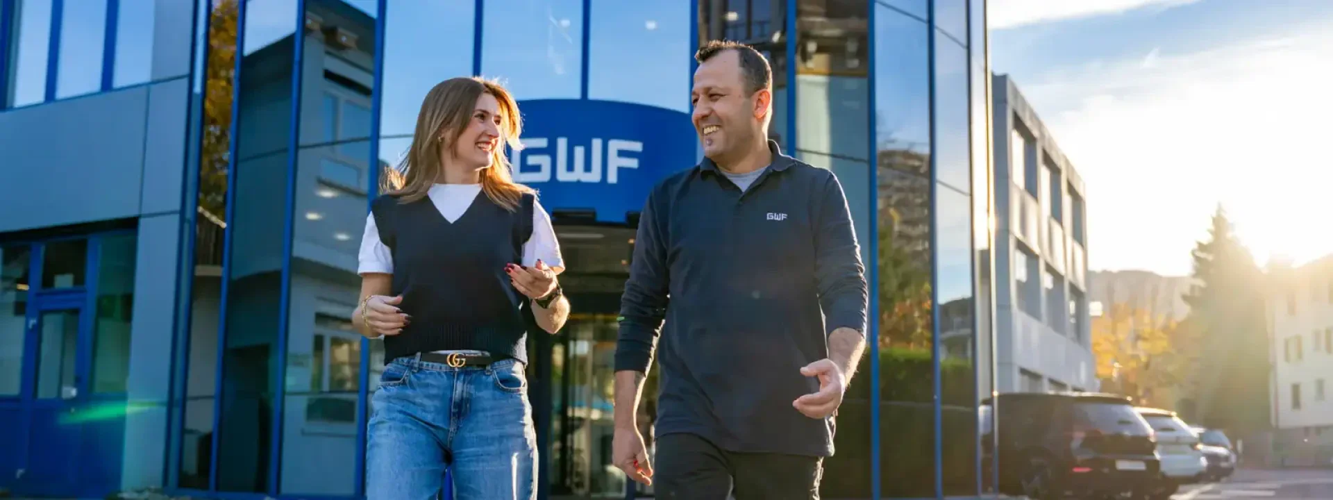 Eine Frau und ein Mann lächeln und unterhalten sich, während sie vor einem modernen gläsernen Bürogebäude spazieren gehen, über dessen Eingang ein Schild mit der Aufschrift GWF prangt. Die Sonne scheint und die Atmosphäre wirkt freundlich und ungezwungen.