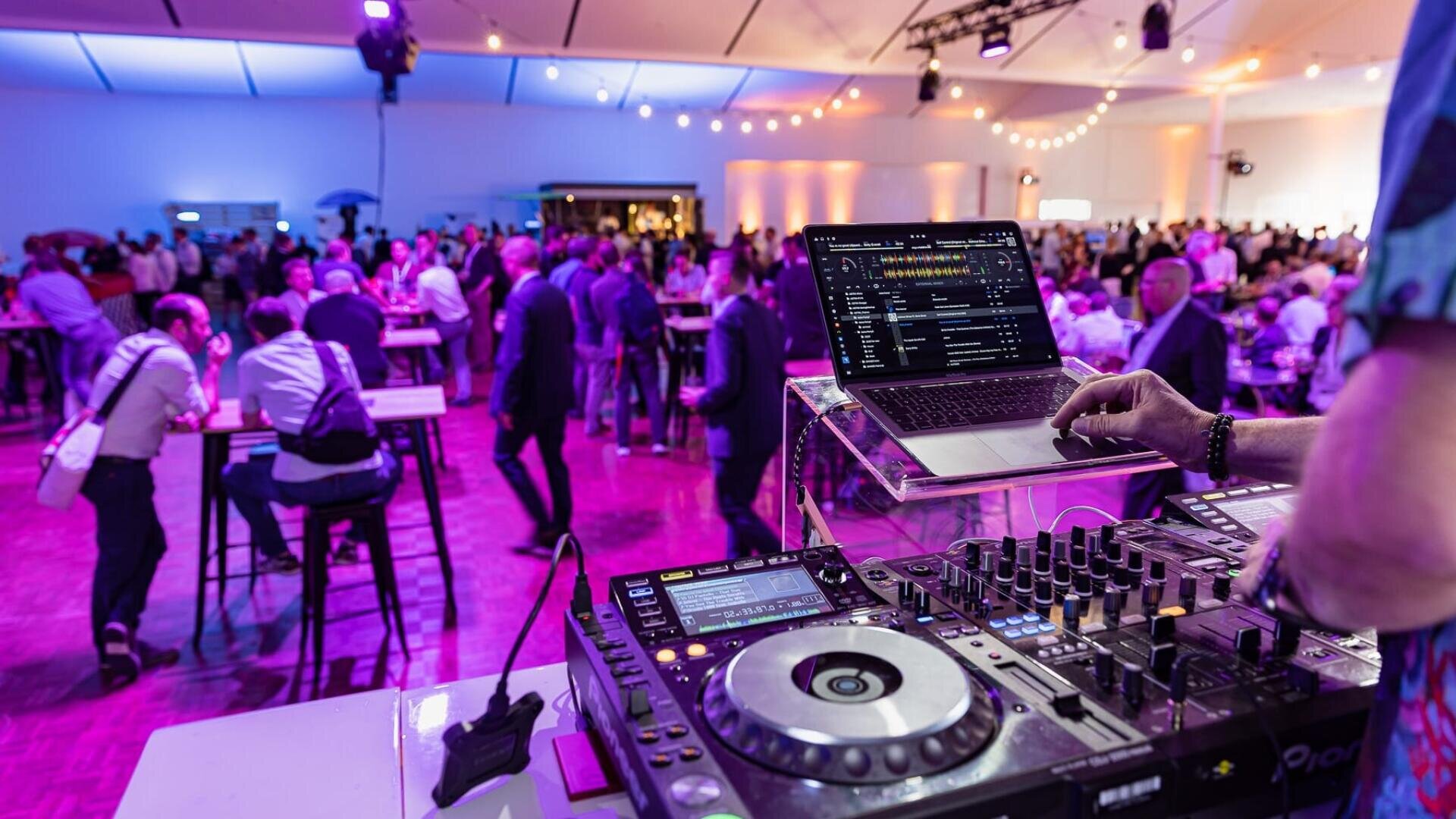 Ein DJ bedient ein Mischpult und einen Laptop bei einer lebhaften Indoor-Veranstaltung mit violetter Beleuchtung, während sich eine Menschenmenge an Stehtischen im Hintergrund unterhält und mischt.
