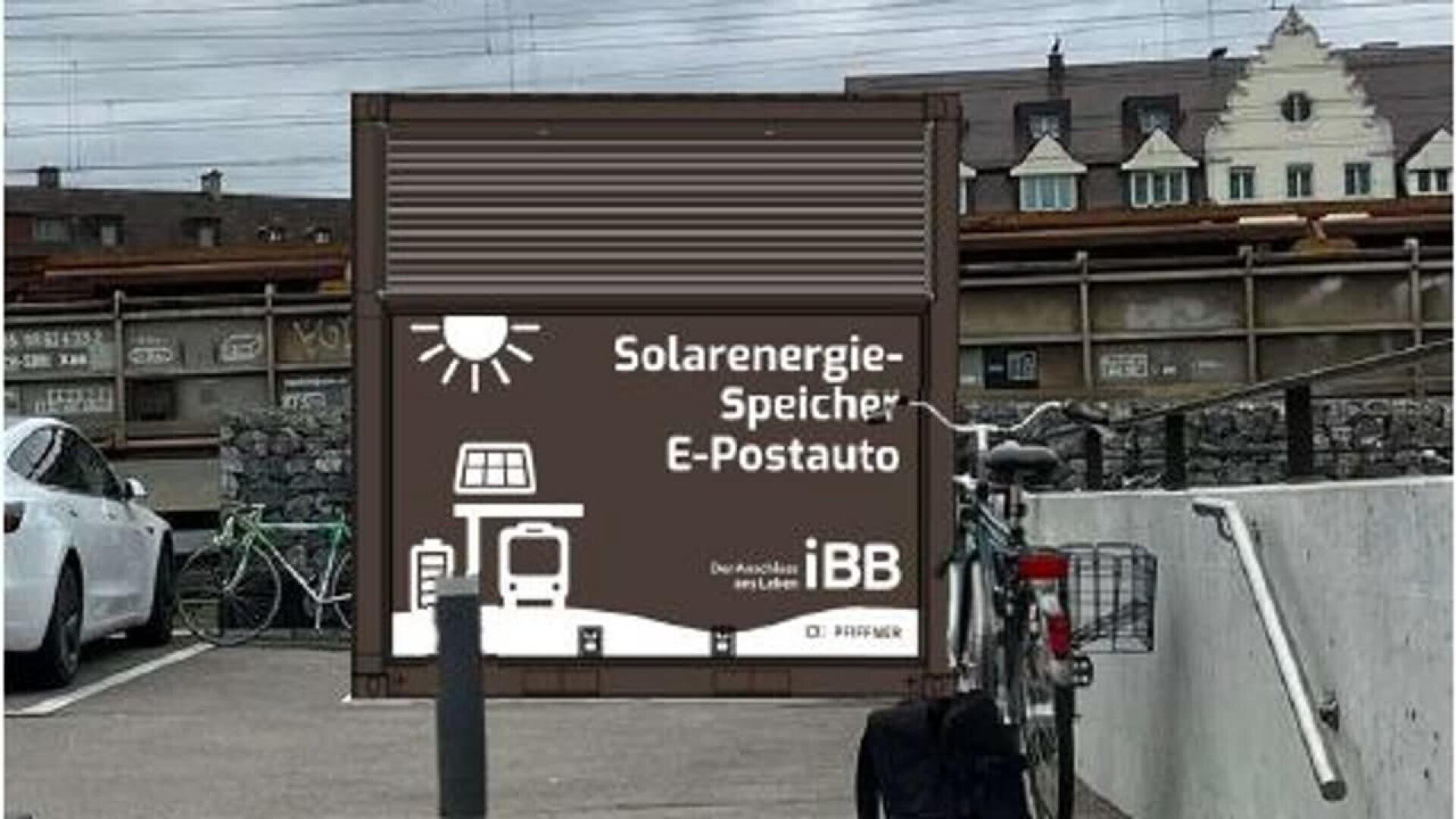 A sign on a building reads Solarenergie-Speicher E-Postauto with icons of the sun, a battery, and an electric bus. Bicycles are parked nearby; a train and historic-style buildings are in the background.