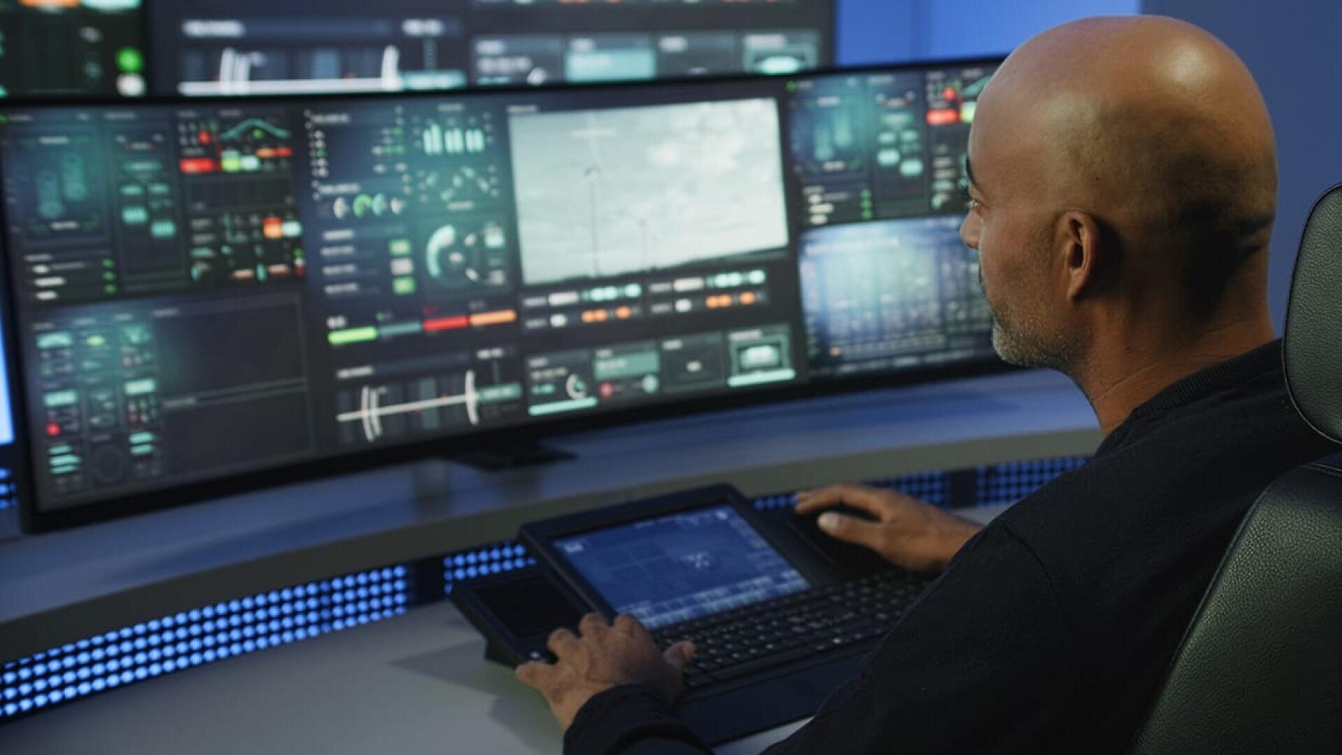 Ein Mann sitzt an einem Schreibtisch und bedient mehrere Computerbildschirme, auf denen komplexe Daten, Radar und Live-Videobilder angezeigt werden. Er scheint ein Hightech-System in einem modernen Kontrollraum zu überwachen oder zu steuern.