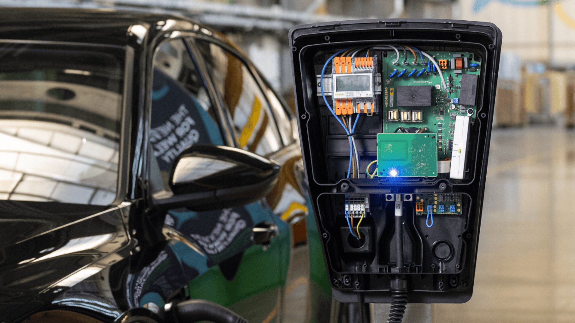 Nahaufnahme einer offenen Ladestation für Elektrofahrzeuge, die die internen Schaltkreise zeigt, mit einem schwarzen Auto, das in der Nähe geparkt ist, in einem gut beleuchteten Industriegebäude.