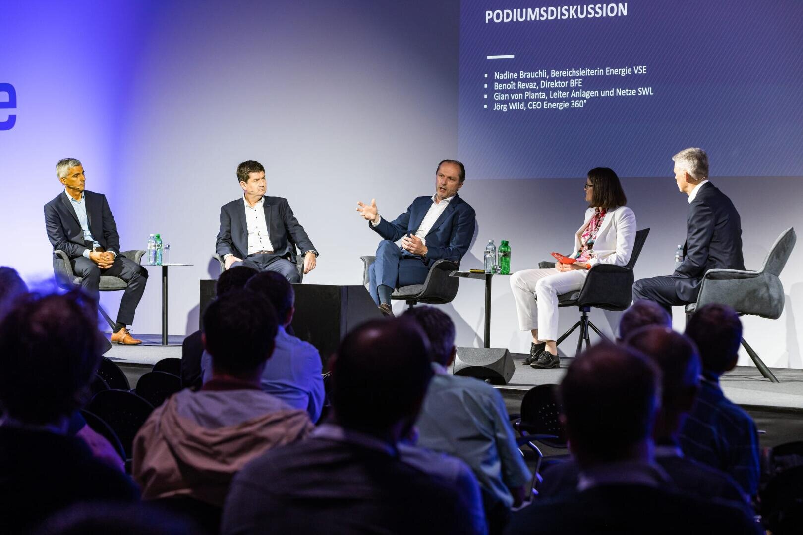 Fünf Personen sitzen auf der Bühne in Sesseln und führen eine Podiumsdiskussion. Das Publikum schaut zu, und auf einer Leinwand hinter ihnen werden Namen und Diskussionsthemen in deutscher Sprache angezeigt. Die Kulisse ist modern, die Beleuchtung professionell.