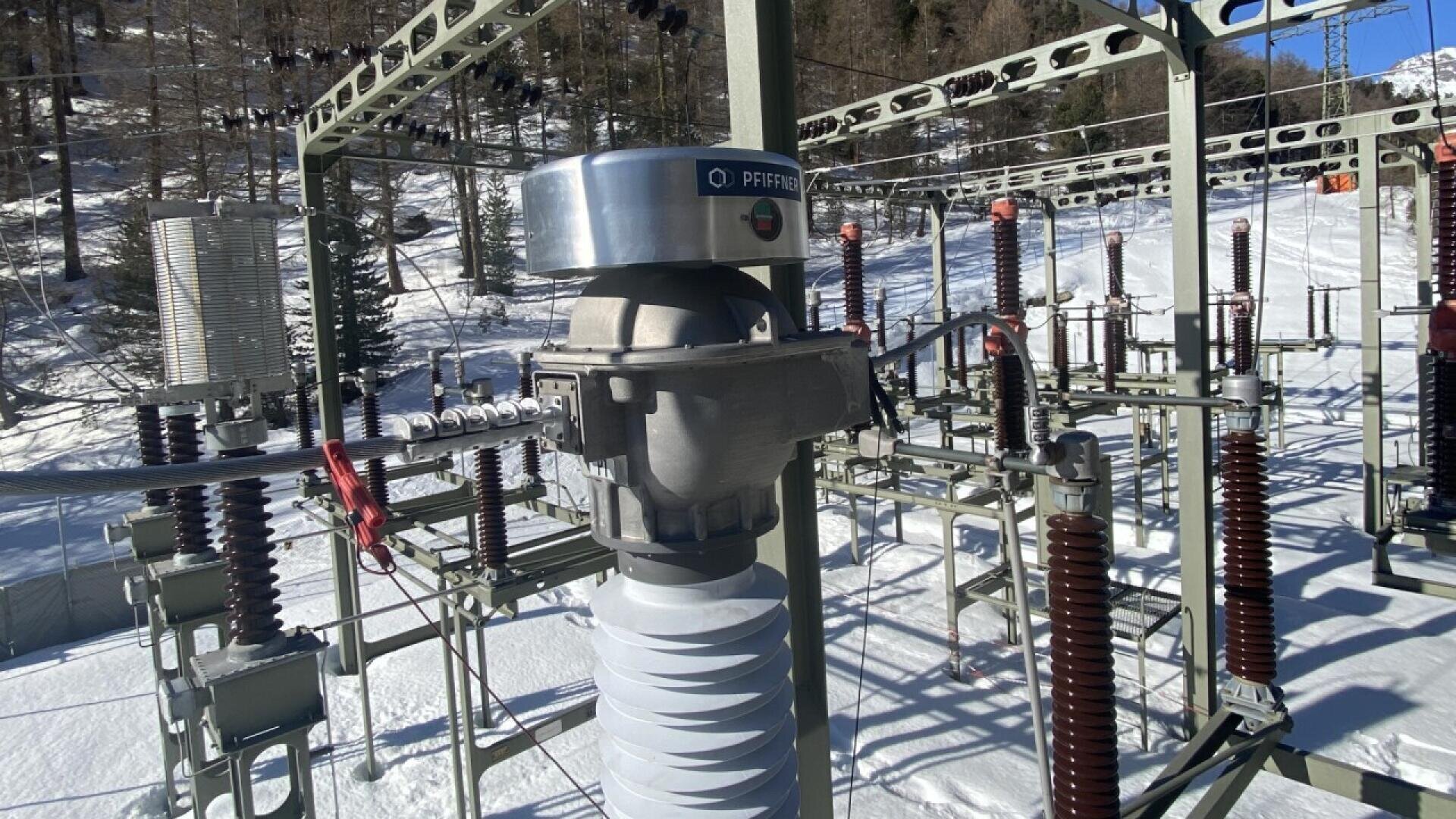 Electrical substation equipment outdoors in a snowy area, featuring various high-voltage insulators and metal structures with mountains and trees visible in the background.
