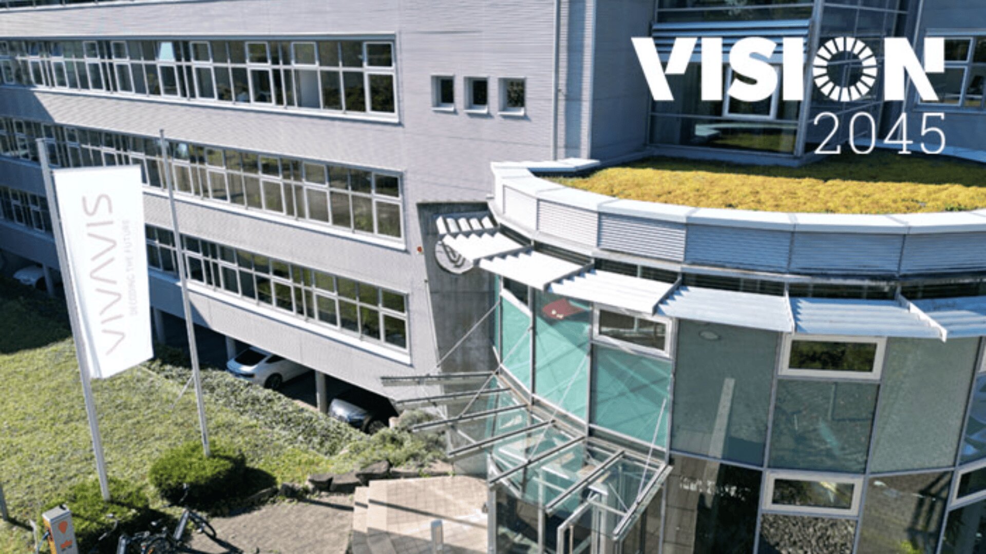 Ein modernes Bürogebäude mit großen Glasfenstern und einem grünen Dach, außen eine weiße Fahne mit VIVAVIS und dem Text VISION 2045 in der oberen rechten Ecke.