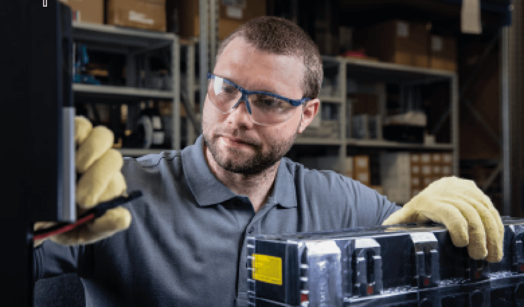 Ein Mann mit Schutzbrille und Handschuhen arbeitet mit elektrischen Geräten in einem Lagerraum und hantiert mit Kabeln und einem Akkupack. Im Hintergrund sind Regale mit Kisten und Werkzeugen zu sehen.
