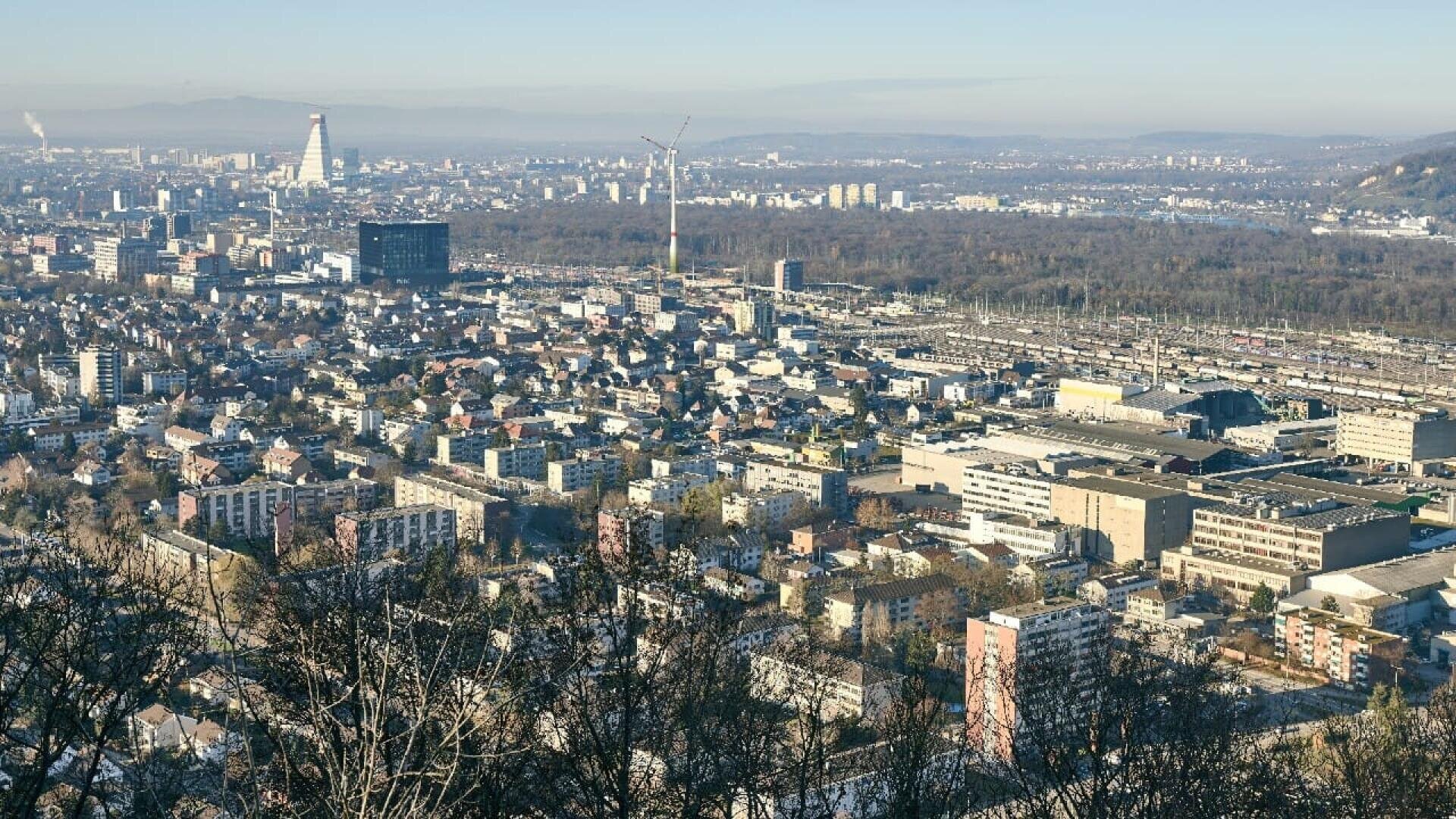 Panoramablick auf eine Stadt mit dichter Bebauung, Industriegebieten und einem markanten Wolkenkratzer in der Ferne, umgeben von Hügeln und Bäumen im Vordergrund unter einem dunstigen Himmel.