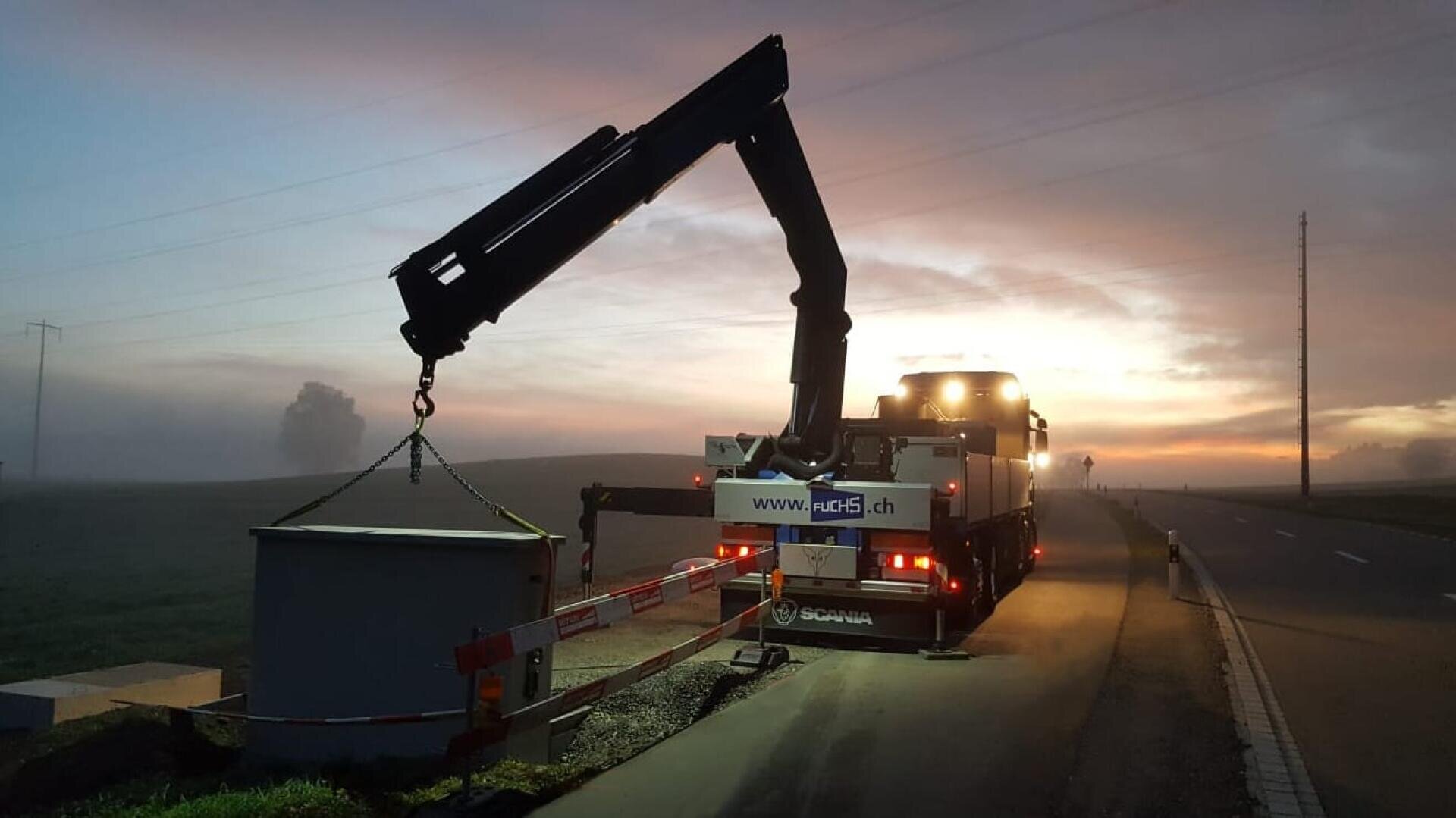 Ein Lastwagen mit einem montierten Kran hebt einen großen grauen Container am Straßenrand in der frühen Morgen- oder Abenddämmerung. Die Szene ist neblig, mit einem Sonnenaufgang oder Sonnenuntergang im Hintergrund und einer ruhigen, ländlichen Straße in der Nähe.