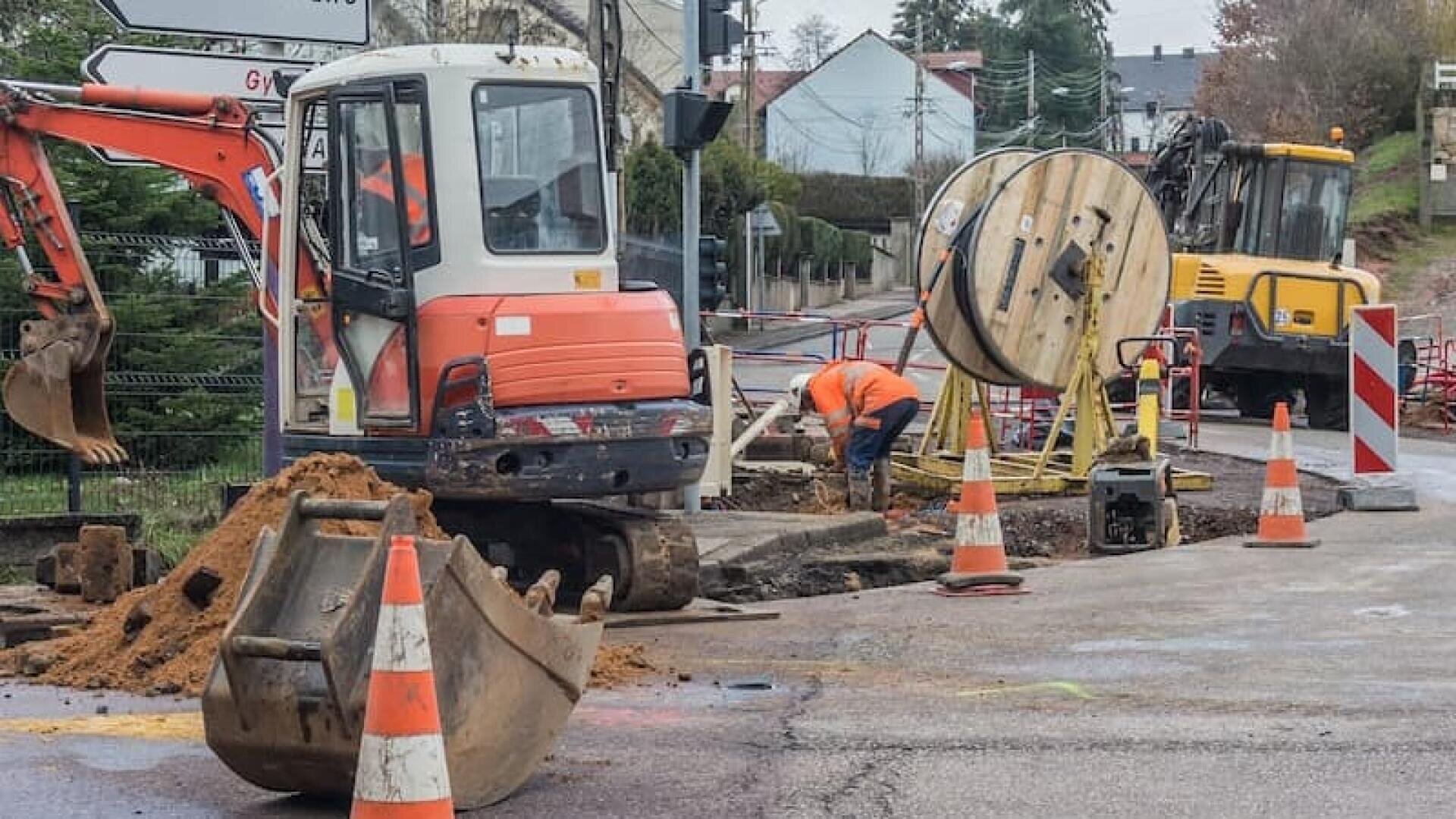 Zwei Bauarbeiter und schwere Maschinen arbeiten auf einer Straße. Verkehrsleitkegel und Absperrungen umgeben einen Graben, in dem Kabel und Geräte zu sehen sind. Im Hintergrund sind Häuser und Bäume zu sehen.