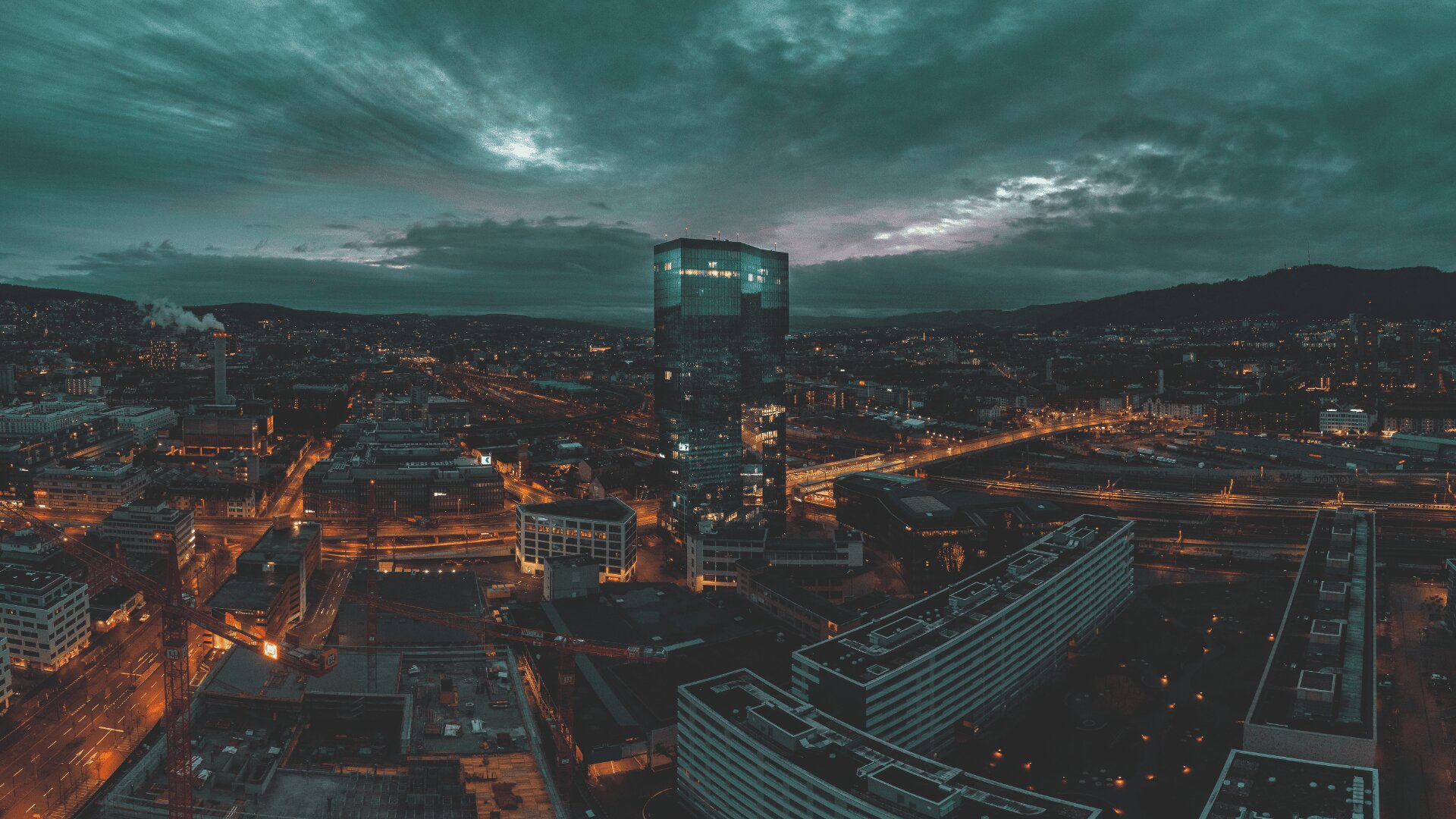 Luftaufnahme einer Stadtlandschaft in der Abenddämmerung mit beleuchteten Straßen, einem hohen gläsernen Wolkenkratzer in der Mitte und dramatischen Wolken über dem Kopf. Gebäude und Straßen werden von Straßenlaternen beleuchtet, wodurch eine lebendige städtische Atmosphäre entsteht.