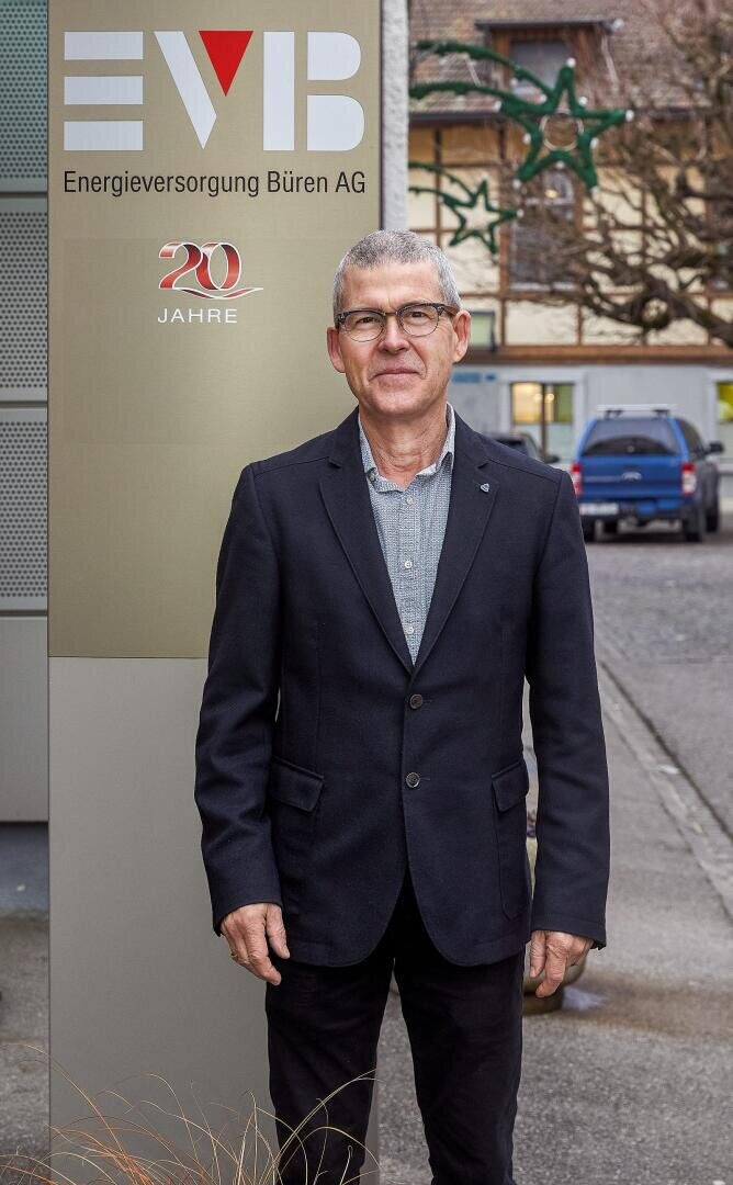 Ein Mann mit dunklem Blazer und Brille steht vor einem Schild der "Energieversorgung Büren AG", auf dem auch "20 Jahre" steht. Im Hintergrund sind Autos und kahle Bäume an einem bewölkten Tag zu sehen.