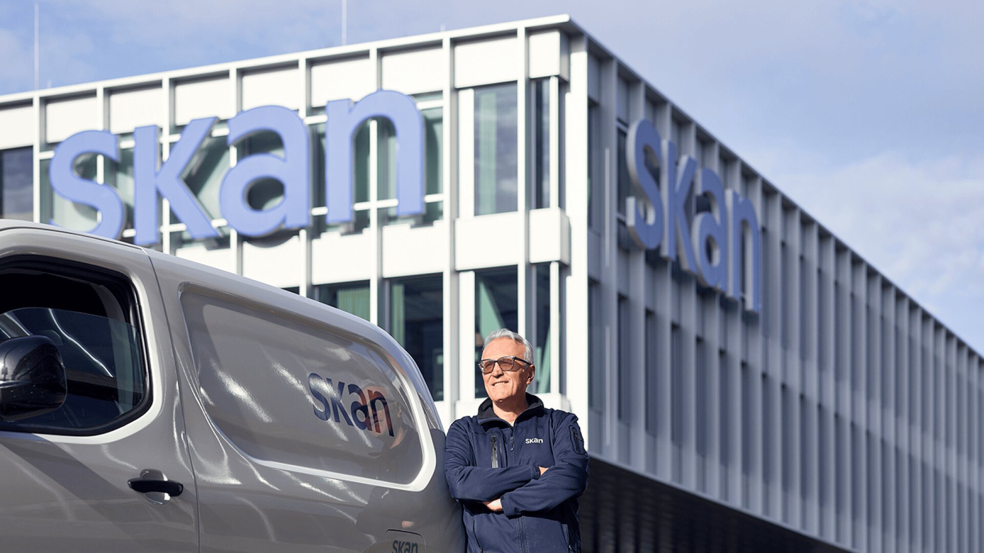 Ein Mann in einer blauen Jacke steht lächelnd mit verschränkten Armen vor einem weißen Lieferwagen und einem modernen Gebäude, die beide das große Logo skan in blauen Buchstaben tragen. Der Himmel ist teilweise bewölkt.