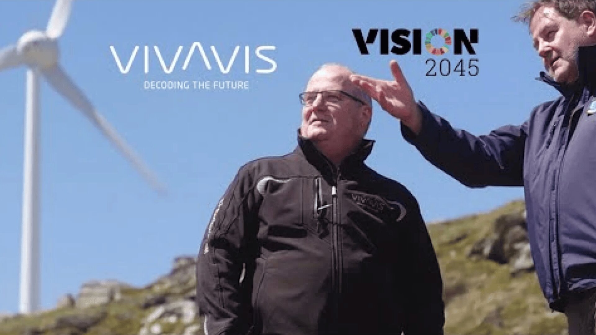 Zwei Männer stehen im Freien in der Nähe einer Windkraftanlage, einer von ihnen gestikuliert in die Ferne. Die Logos von VIVAVIS Decoding the Future und VISION 2045 erscheinen am Himmel über ihnen. Im Hintergrund sind felsige Hügel und blauer Himmel zu sehen.