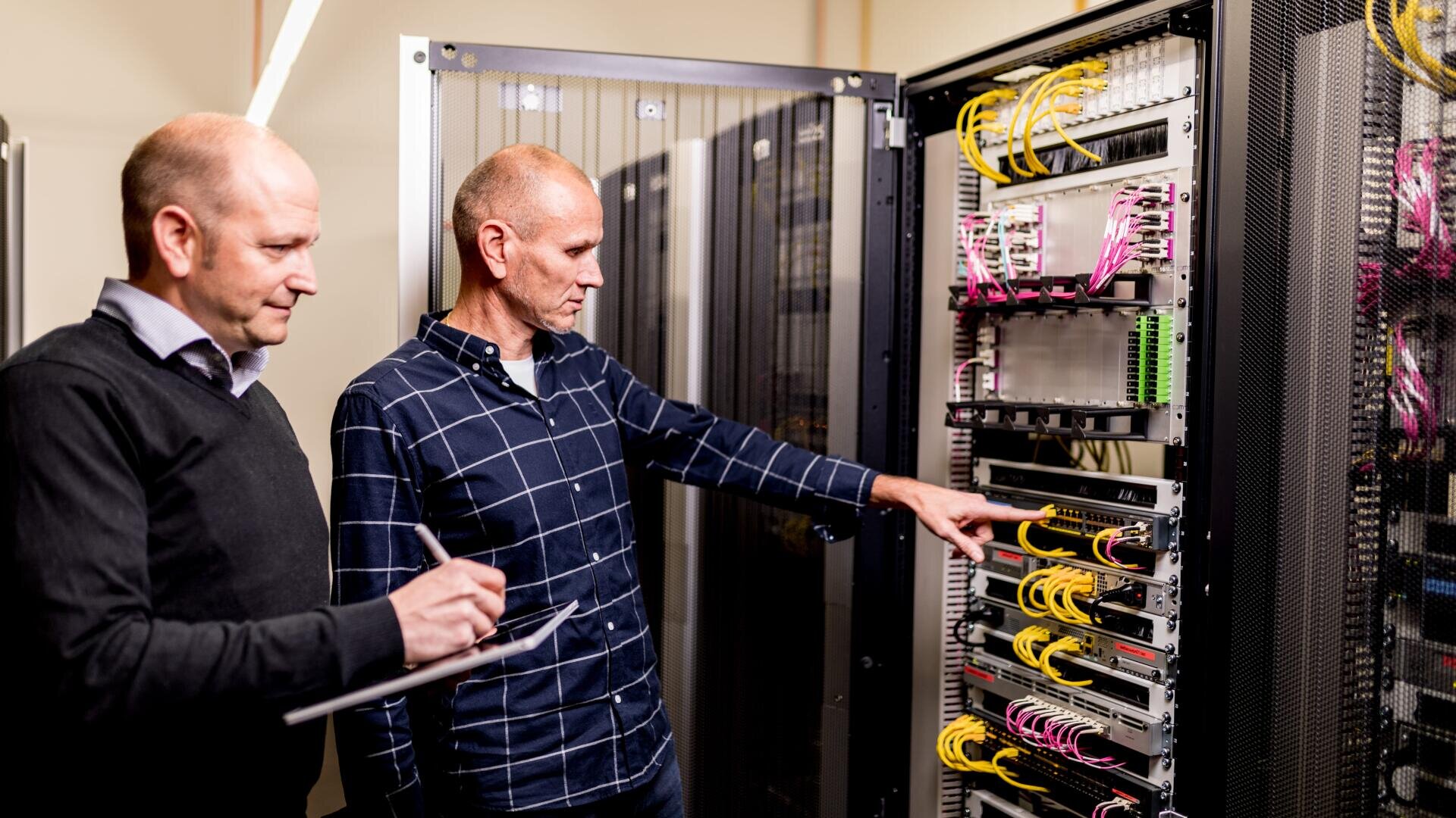 Zwei Männer stehen in einem Serverraum; einer deutet auf Netzwerkgeräte in einem Serverschrank, während der andere ein Klemmbrett hält und beobachtet. Die sichtbaren Kabel sind geordnet und an verschiedene Geräte angeschlossen.