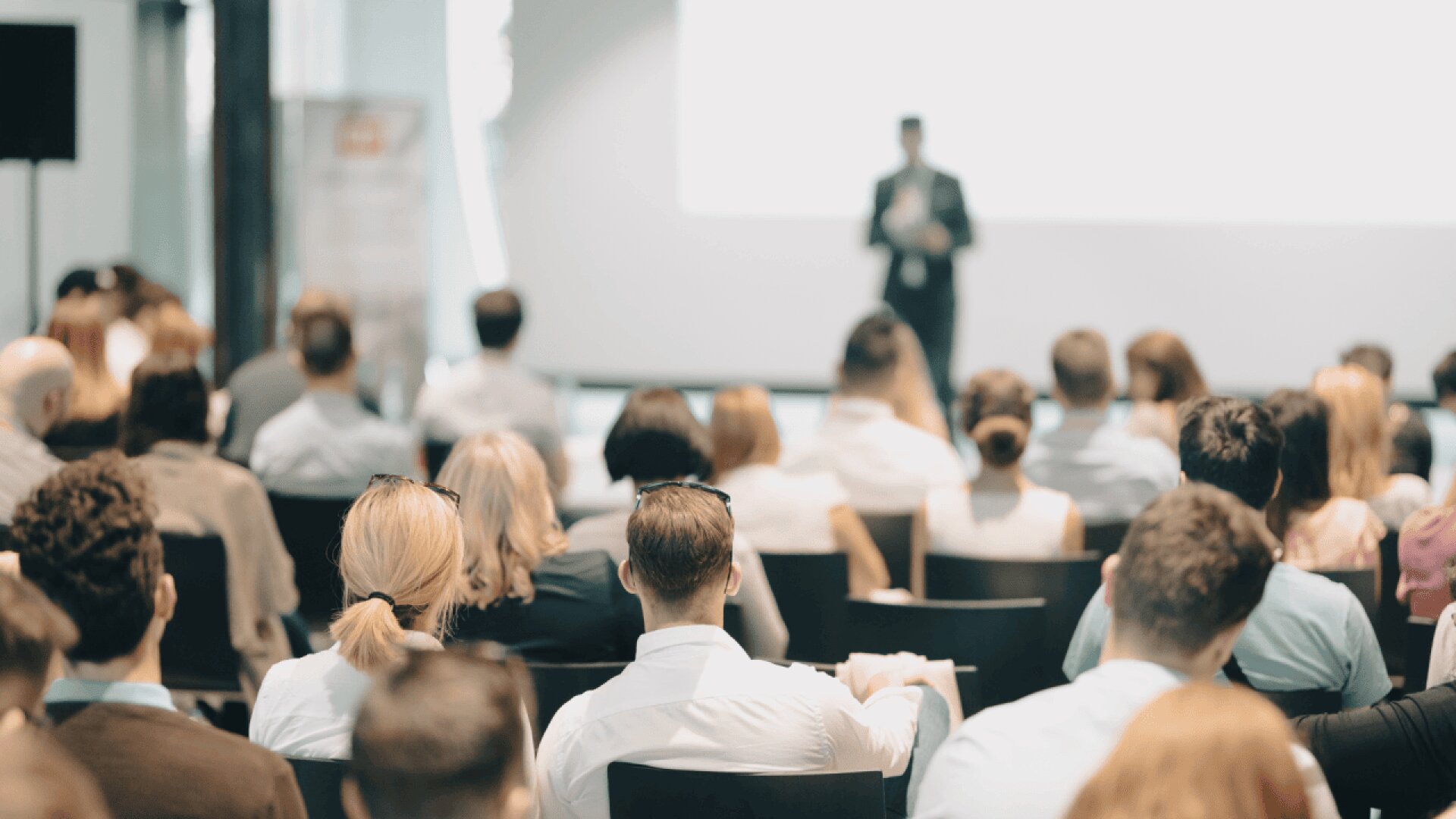A group of people sit in rows, facing a blurred speaker standing in front of a large screen at a conference or seminar in a bright, modern room.