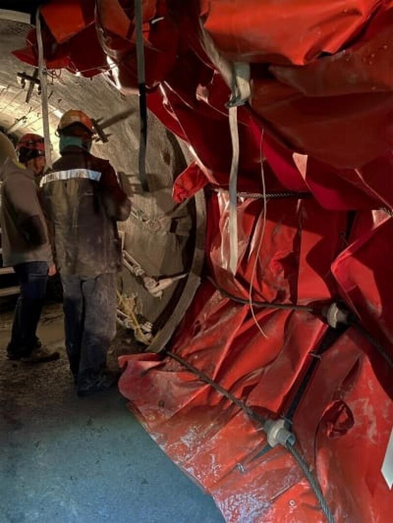 Zwei Arbeiter in Schutzkleidung stehen in einem Tunnel in der Nähe einer mit rotem Schutzmaterial ausgekleideten Wand, die mit Bolzen und Gurten gesichert ist. Der Tunnel ist schwach beleuchtet, und im Hintergrund ist Baumaterial zu sehen.