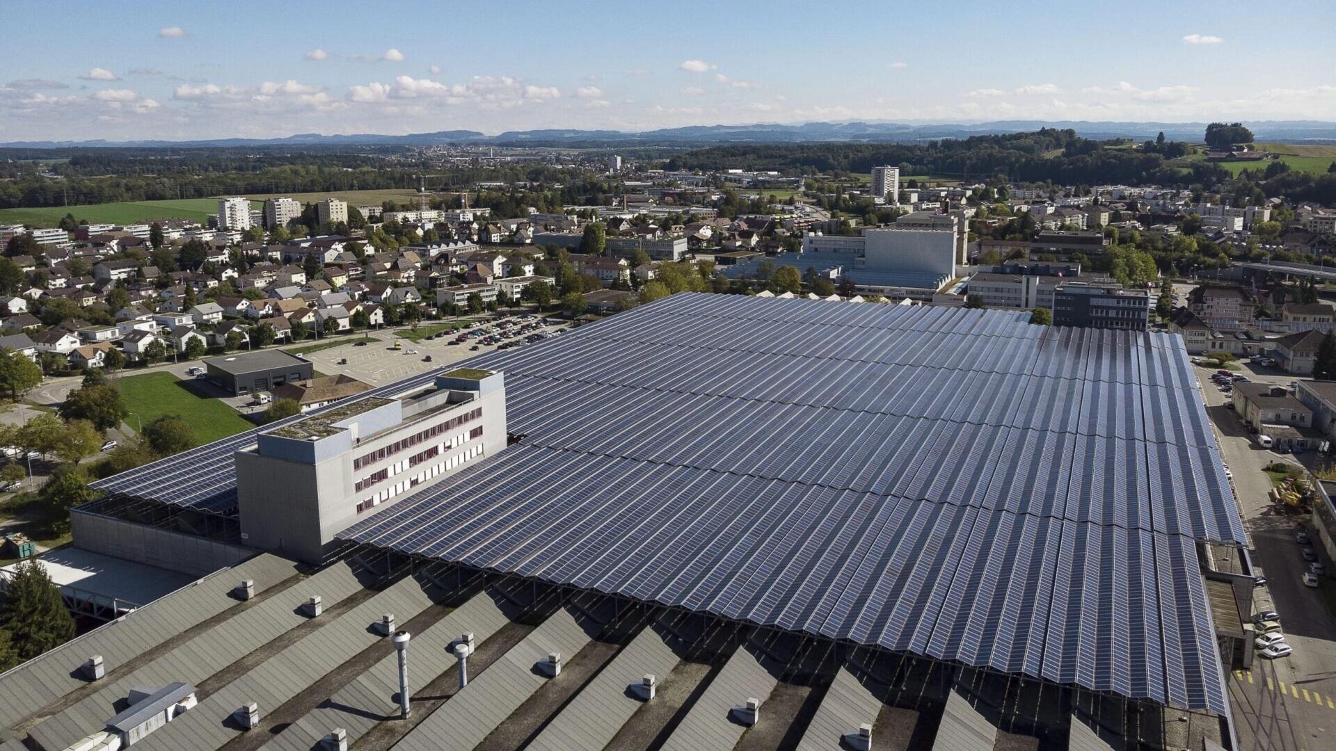 Ein großes Industriegebäude mit einem mit Solarzellen bedeckten Dach ist in einer Luftaufnahme zu sehen, umgeben von einem Wohngebiet, grünen Feldern und entfernten Hügeln unter einem teilweise bewölkten Himmel.