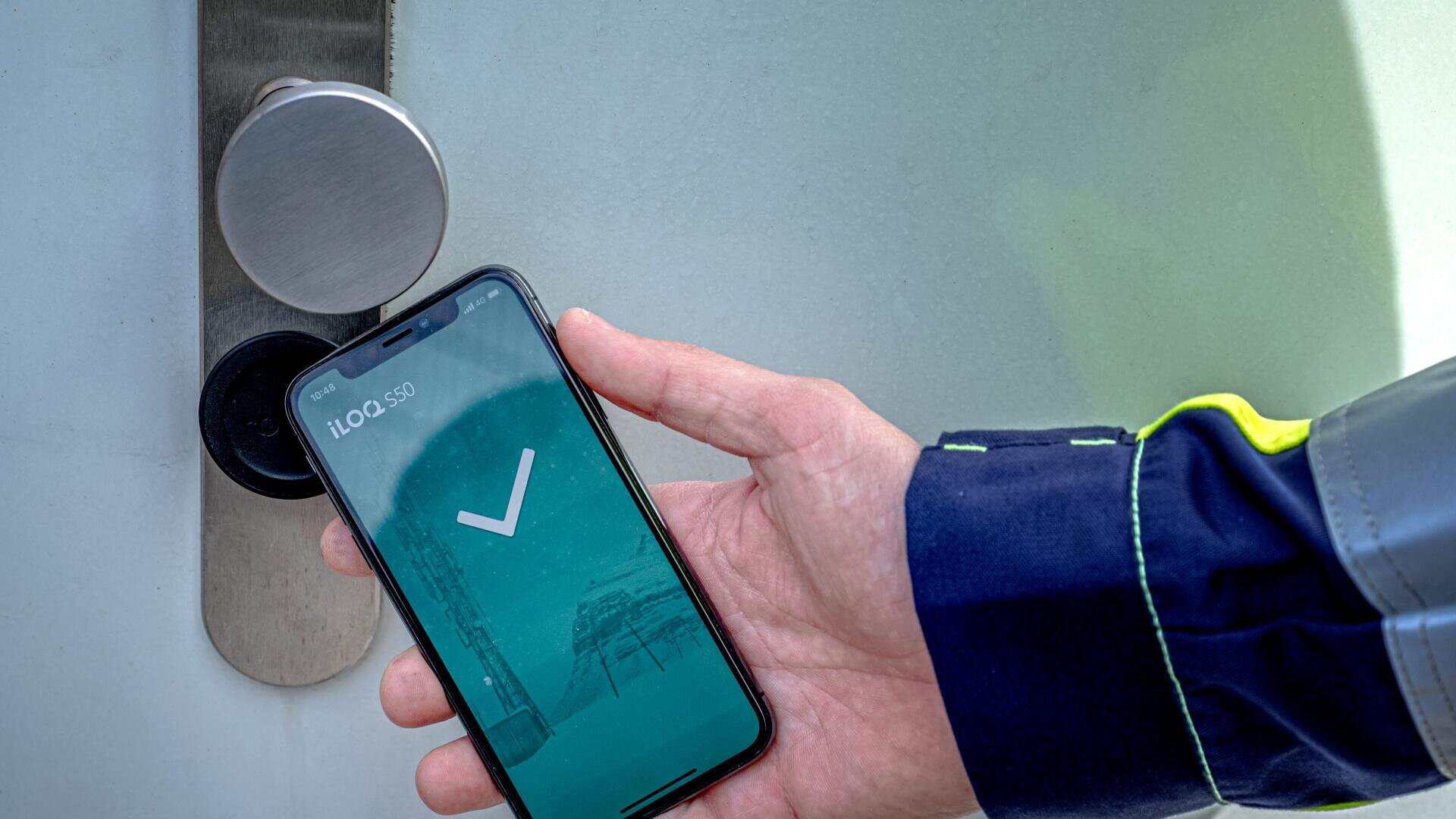 A person holds a smartphone showing a checkmark screen next to a modern electronic door lock, preparing to unlock the door. The person is wearing a blue and yellow jacket.