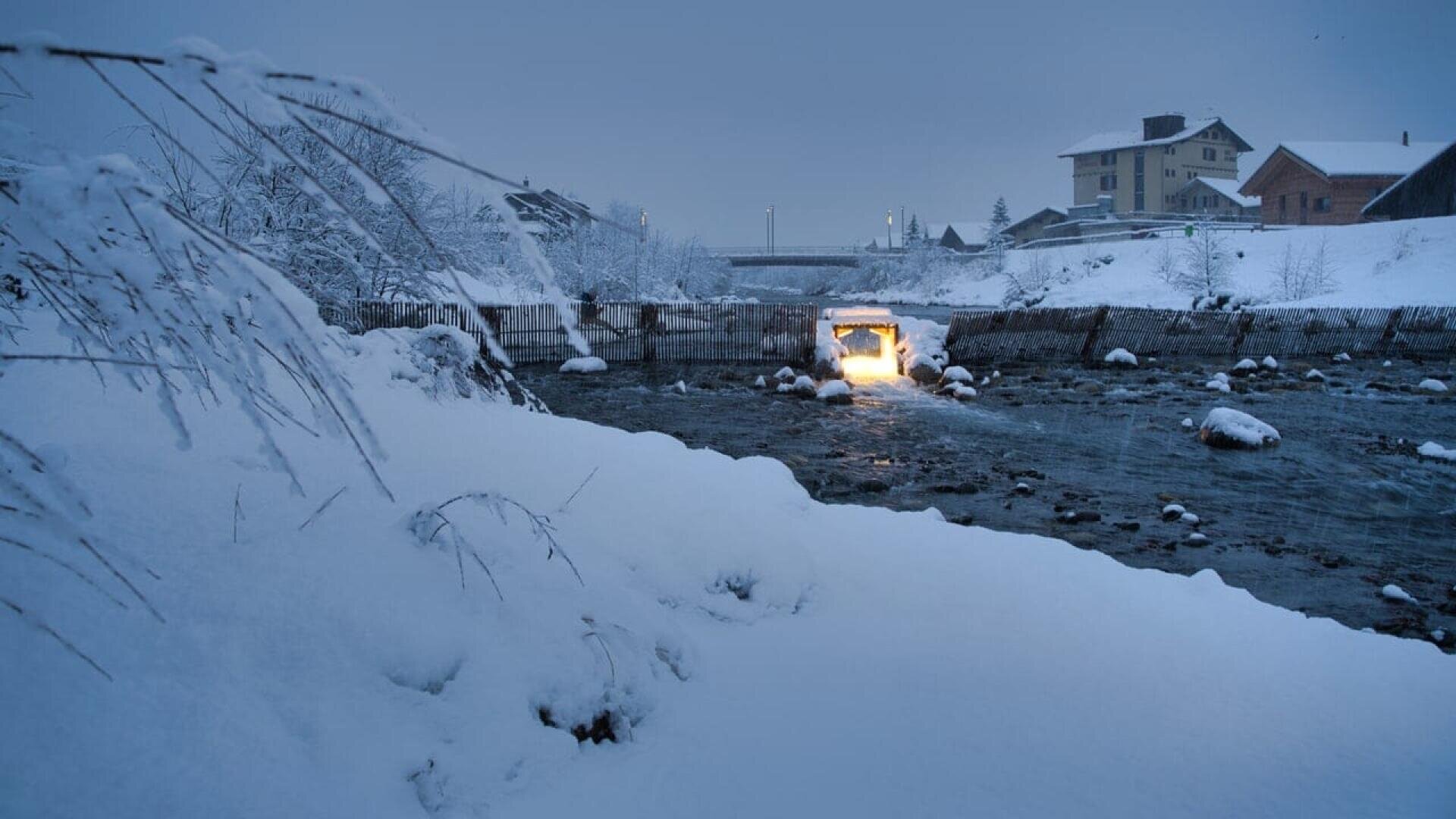A snowy riverbank at dusk with a stream flowing under a bridge. Buildings and trees are covered in snow, and a warm light glows from an opening beneath the bridge, illuminating the cold scene.