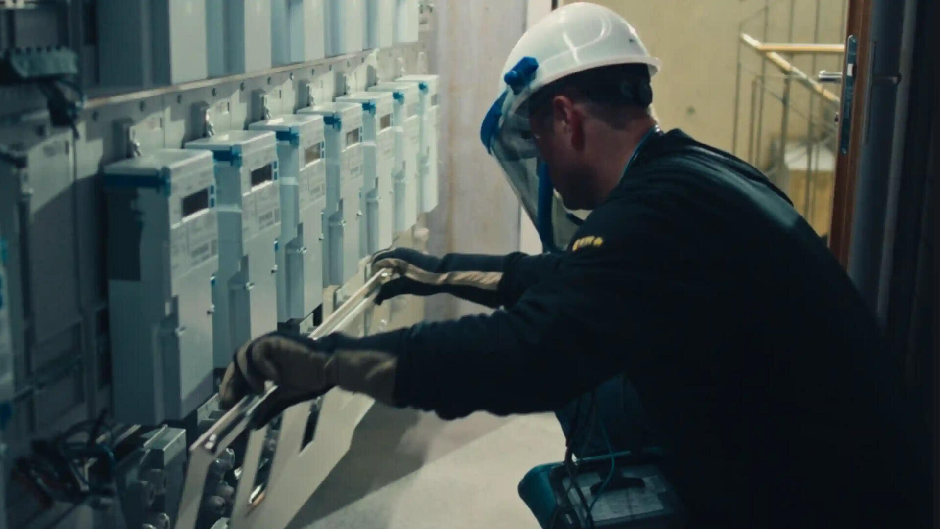 Ein Elektriker, der einen weißen Schutzhelm, eine Schutzbrille, Handschuhe und dunkle Kleidung trägt, arbeitet an einer elektrischen Schalttafel mit mehreren Zählern oder Geräten in einem Gebäude.