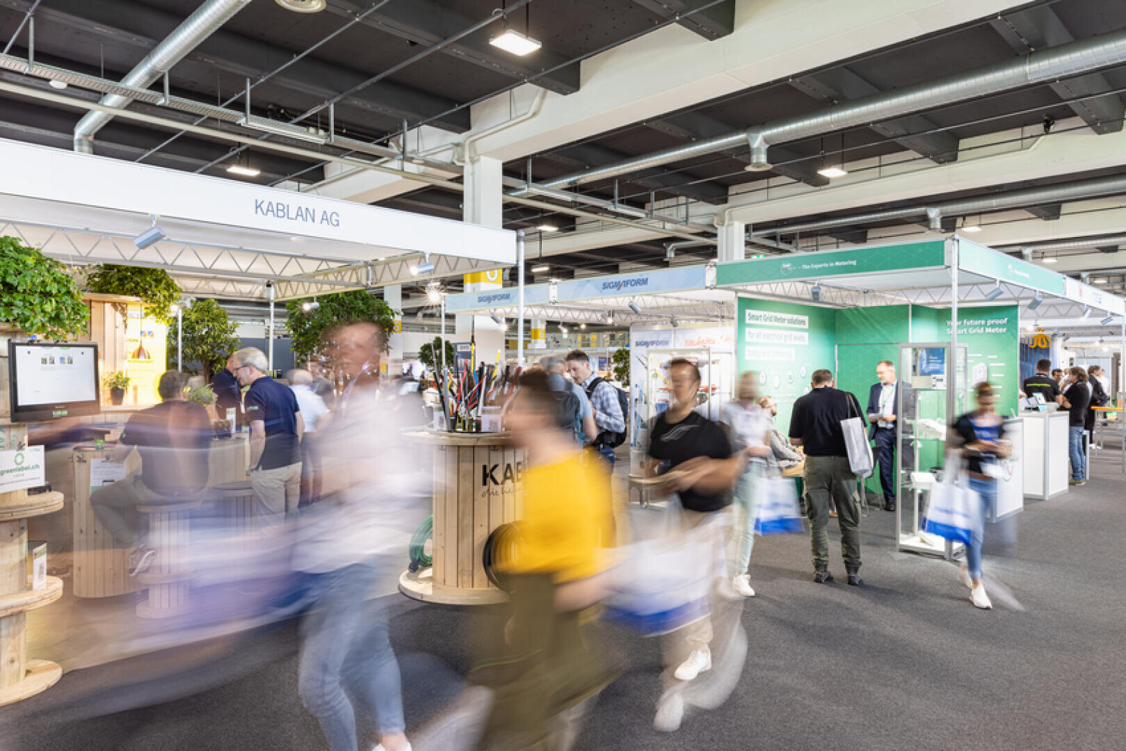 Eine gut besuchte Messe in einer Halle, auf der Menschen an verschiedenen Messeständen vorbeigehen, darunter auch an einem Stand der KABLAN AG. Einige Besucher sind in Bewegung und wirken verschwommen, sie tragen Taschen, während andere an den Ständen mit den Ausstellern ins Gespräch kommen. Die Atmosphäre ist lebhaft und geschäftig.