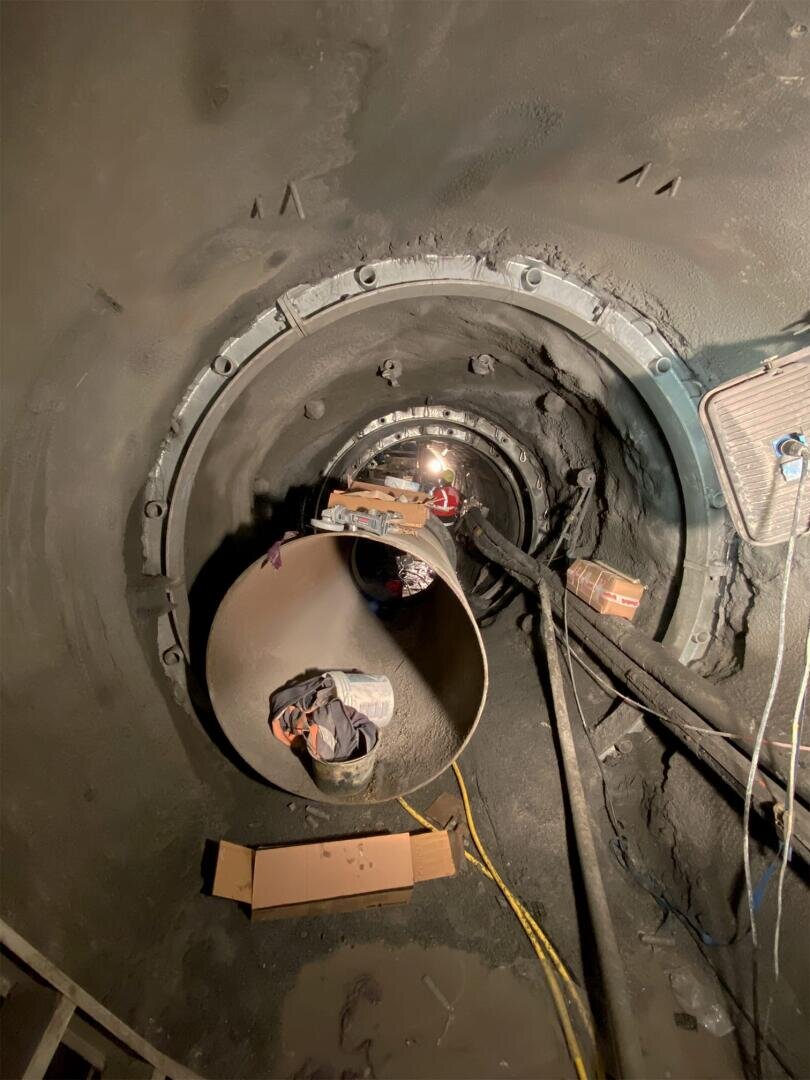 Blick in einen großen, zylindrischen Tunnel mit Geräten, Kabeln und Baumaterialien. Im Vordergrund sind ein Rohr und eine Pappschachtel zu sehen. Ein helles Licht beleuchtet die Länge des Tunnels.