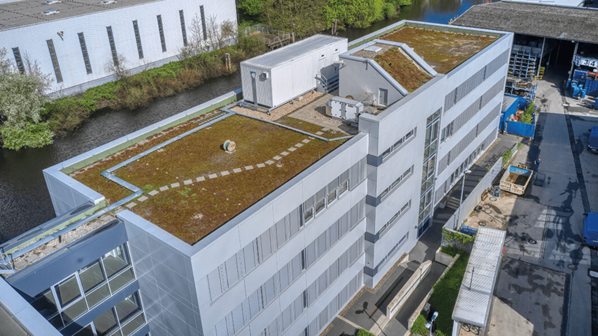 Luftaufnahme eines modernen grauen Gebäudes mit begrüntem Dach, Wegen und HLK-Anlagen, umgeben von anderen Gebäuden, Wasser, Bäumen und einem Industriegebiet.