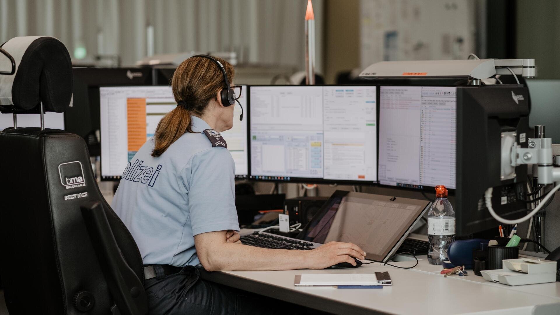 Eine Frau in Polizeiuniform sitzt an einem Schreibtisch mit mehreren Computermonitoren, trägt ein Headset und schaut auf die Bildschirme. Der Arbeitsplatz ist mit Büromaterial, einer Wasserflasche und einem Notizblock ausgestattet.