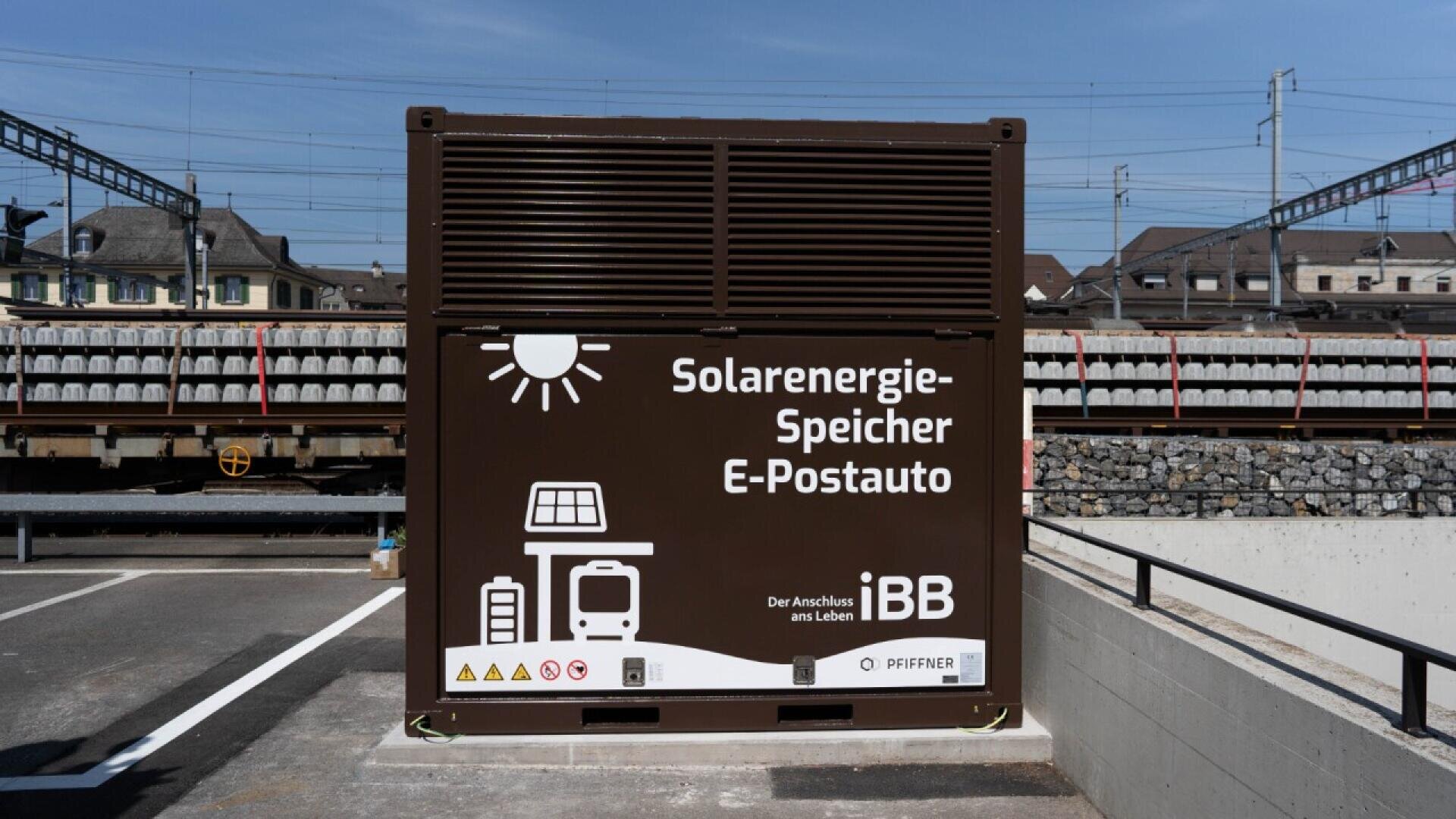 A brown solar energy storage unit labeled Solarenergie-Speicher E-Postauto stands in a parking lot near train tracks, with illustrated icons of the sun, solar panels, a bus, and a car.