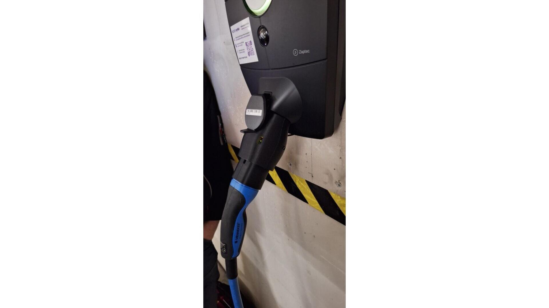 Nahaufnahme einer Ladestation für Elektrofahrzeuge mit einem schwarz-blauen Ladestecker, der in die Steckdose eingesteckt und an einer mit gelben und schwarzen Warnstreifen versehenen Wand angebracht ist.