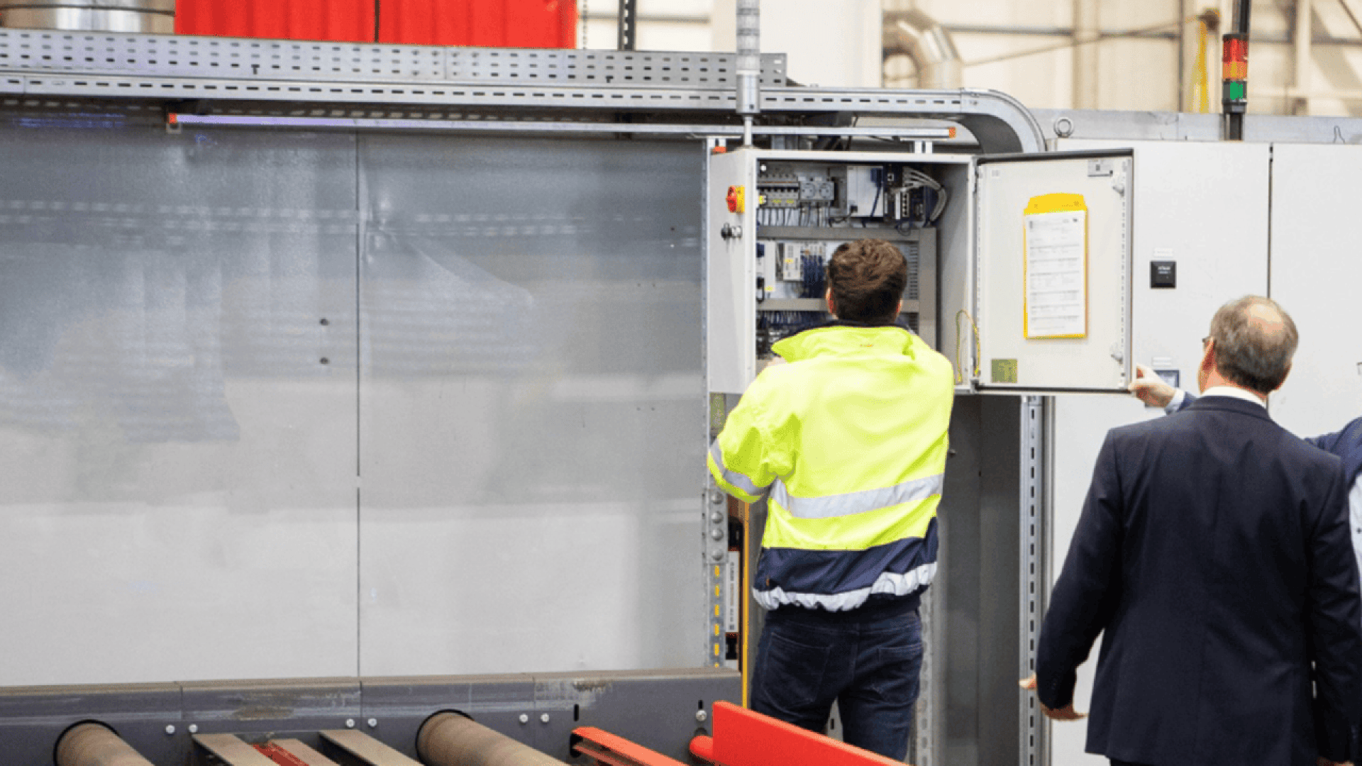 A worker in a high-visibility jacket examines electrical controls inside an open panel, while another person in a suit stands nearby and points at the equipment in an industrial setting.