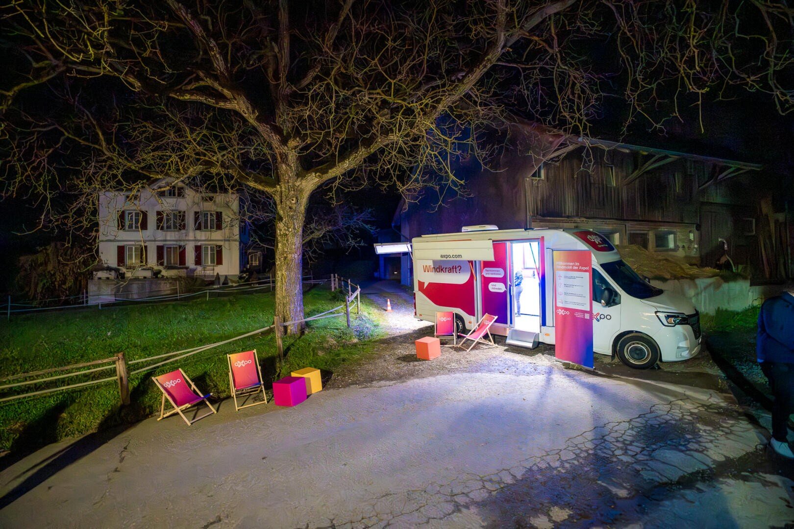 Ein hell erleuchteter Lieferwagen mit der Wirkungsperle auf der Seite ist nachts in der Nähe eines großen Baumes geparkt. Draußen sind Liegestühle und bunte Würfel aufgestellt, und im Hintergrund ist ein Bauernhaus zu sehen.