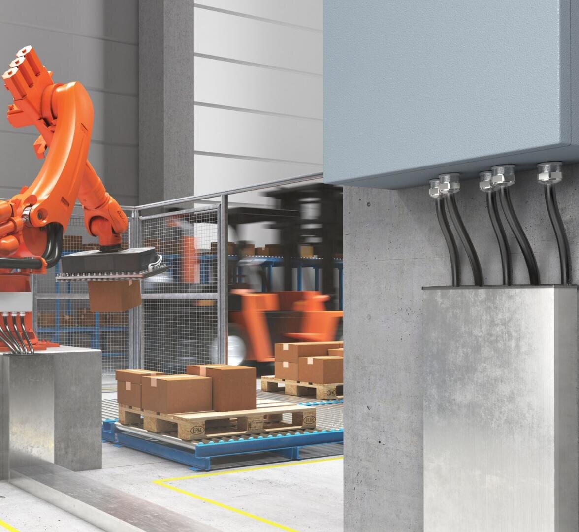 Ein Roboterarm bewegt in einer Fabrik braune Pappkartons auf Holzpaletten. In der Szene sind Metallzäune, Industrieanlagen und elektrische Kabel zu sehen, die an einem an der Wand montierten Schaltkasten befestigt sind.