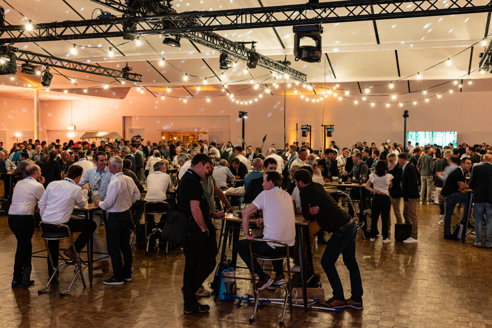 Eine große Gruppe von Menschen versammelt sich in einer geräumigen, gut beleuchteten Halle, plaudert und trifft sich an Stehtischen. Lichterketten und Deckenleuchten schaffen eine warme, festliche Atmosphäre.