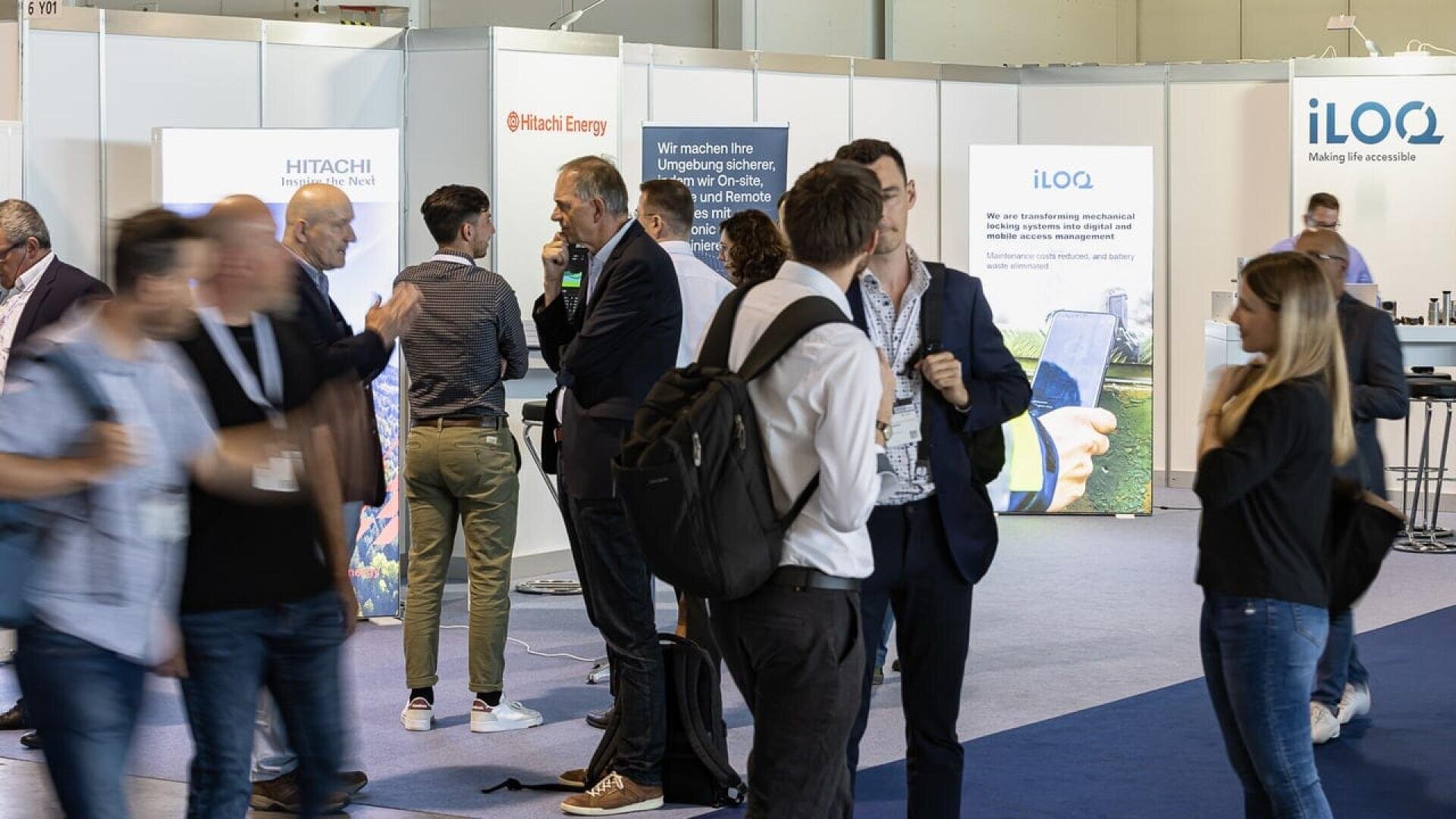 A group of people at a business expo or networking event, some talking in pairs or groups, with informational booths and banners for companies like Hitachi Energy and iLOQ in the background.