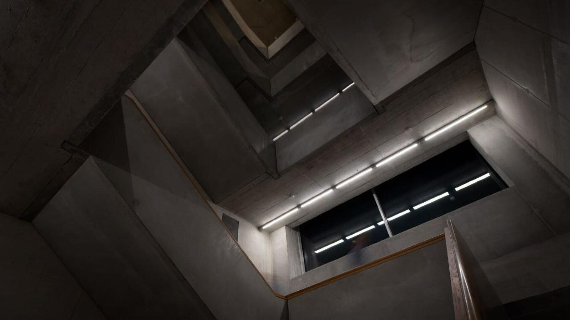 Ein Blick durch ein modernes Betontreppenhaus mit kantigen Wänden, Lichtbändern und einem Fenster, das etwas Licht von oben hereinlässt, schafft eine geometrische, minimalistische Architekturkulisse.