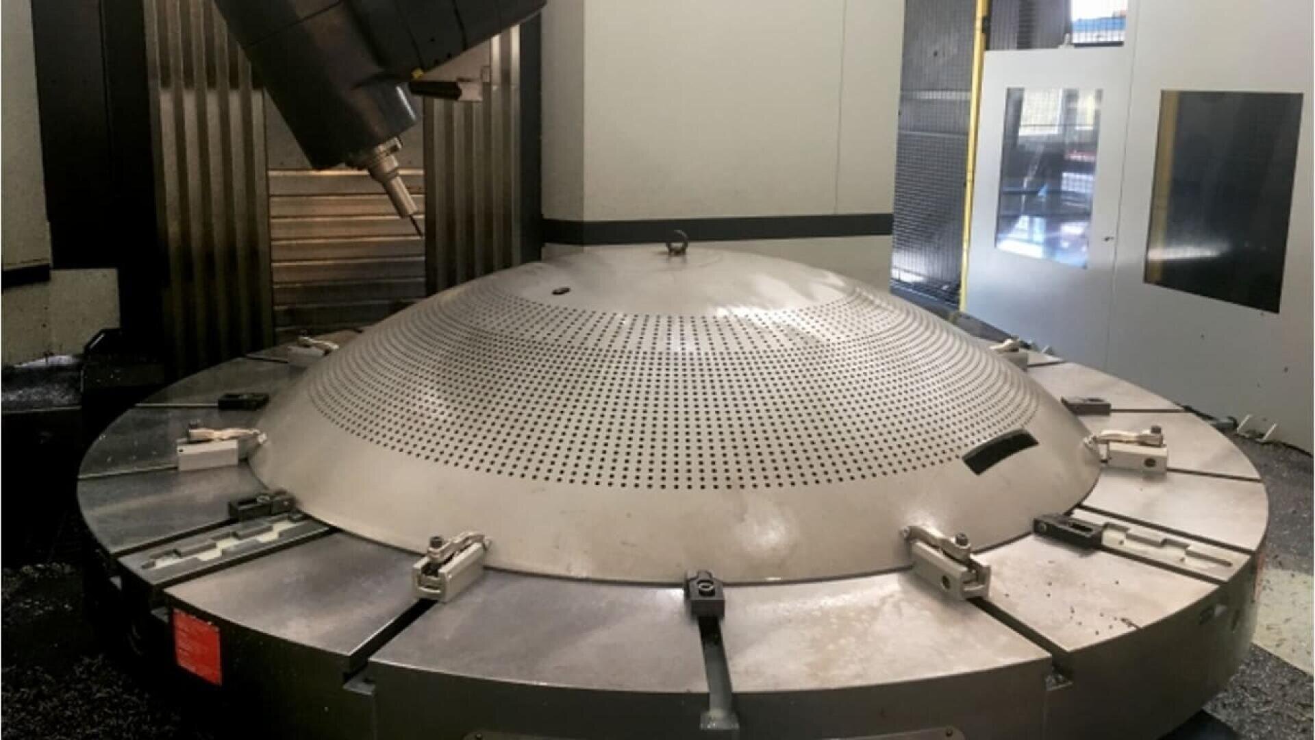 A large, circular metal workpiece with many small holes is clamped onto a CNC machine table, with a milling head positioned above it for precision machining inside an industrial workshop.