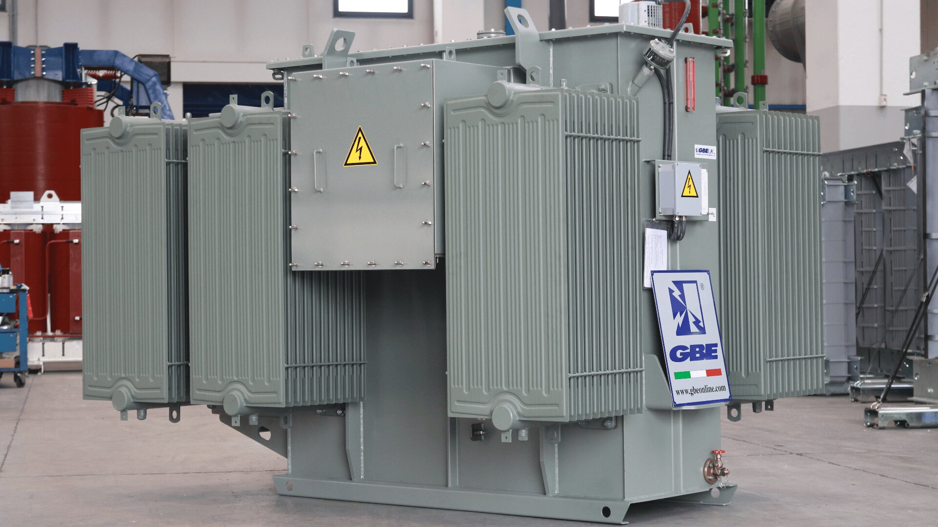 A large industrial electrical transformer with cooling fins and warning signs stands indoors on a concrete floor. The transformer is grey with a GBE label and various connectors and components visible.