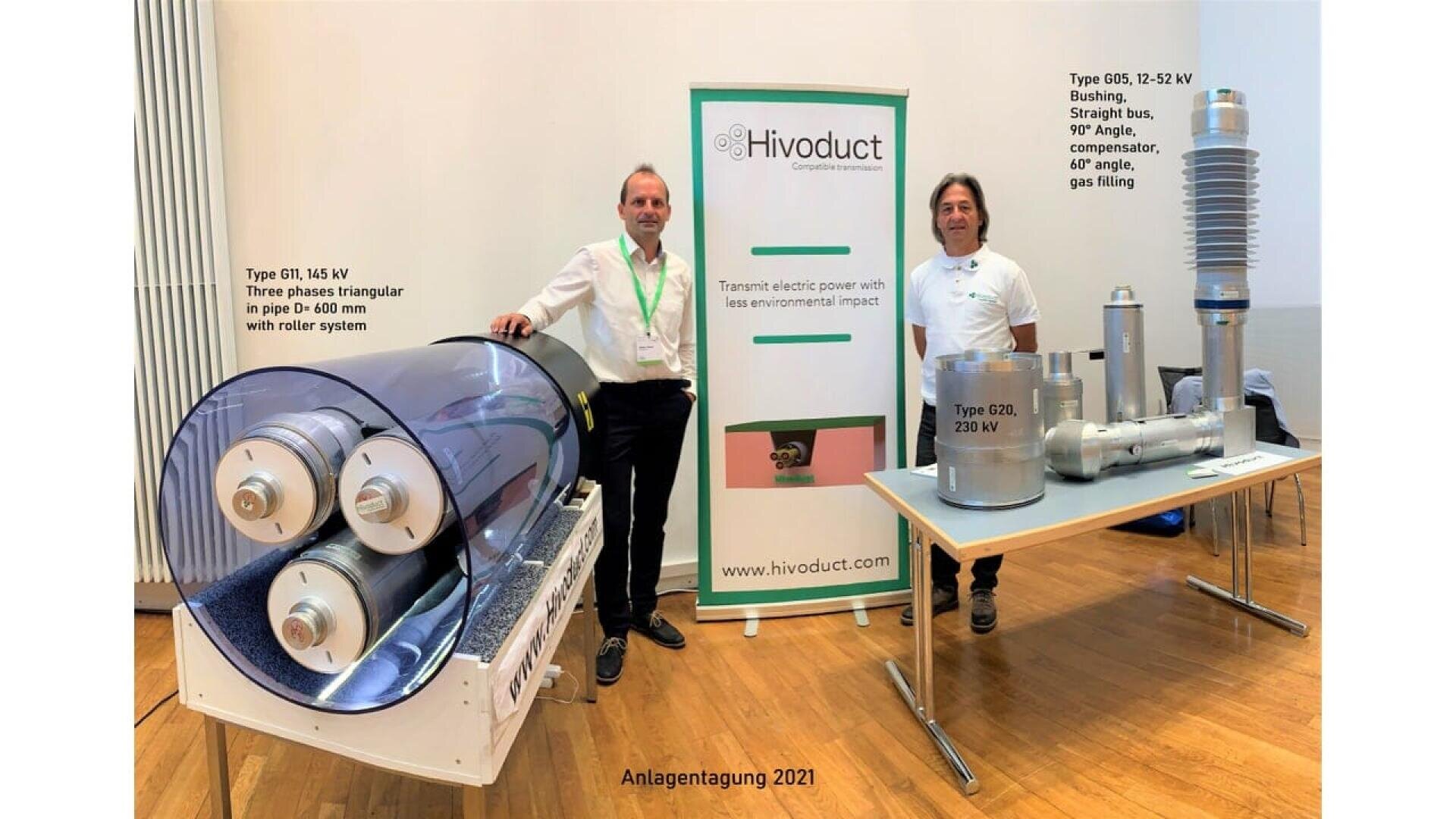 Two men stand beside a Hivoduct display at a conference. Two types of electrical components are shown: a large cylindrical unit on the left in a transparent casing, and several metallic devices on the right. Banner reads “Transmit electric power with less environmental impact.”.