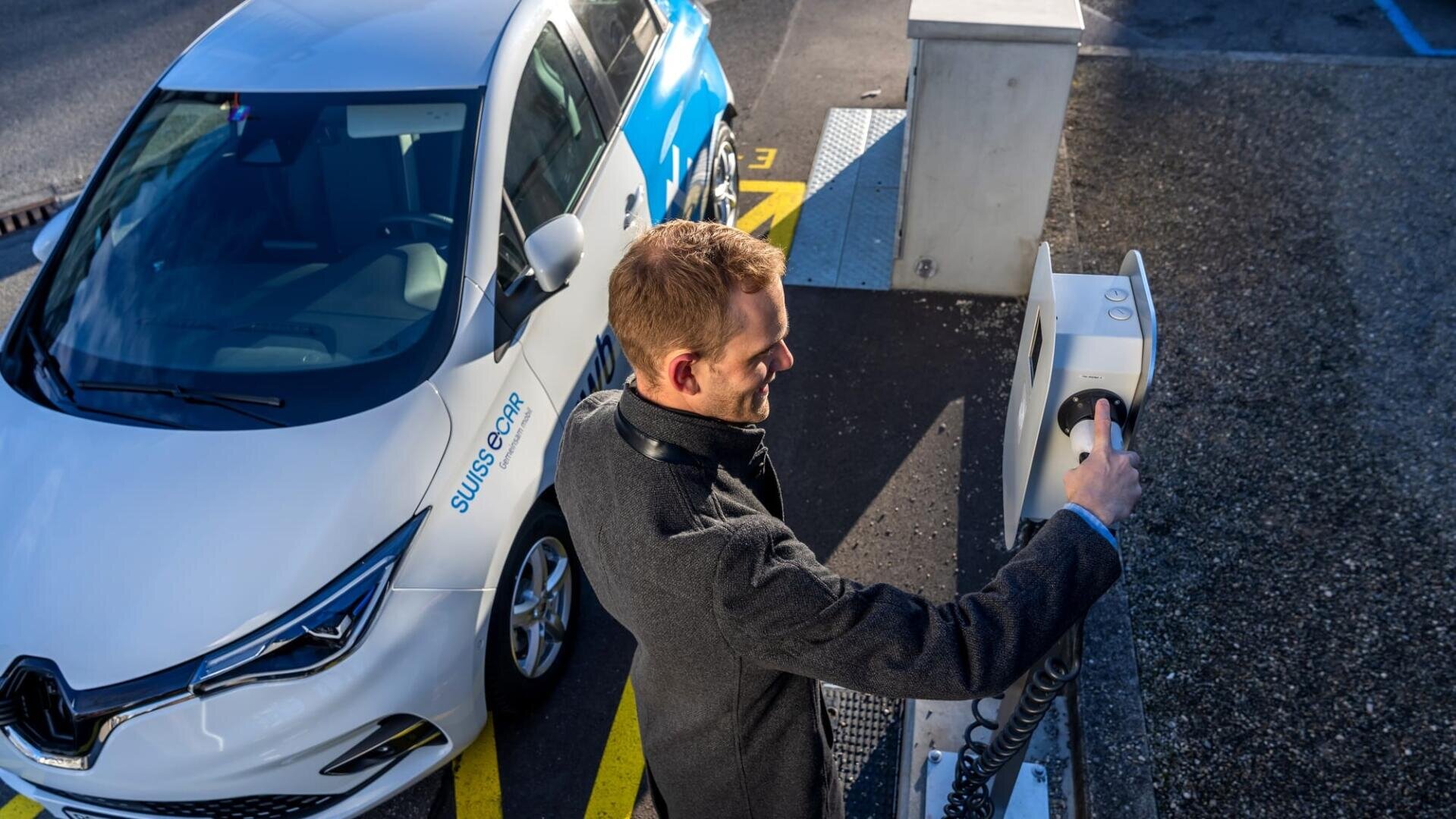 Ein Mann in einem dunklen Mantel schliesst an einer Ladestation im Freien ein Ladekabel an ein weisses Elektroauto mit SWISSCOM-Branding an.