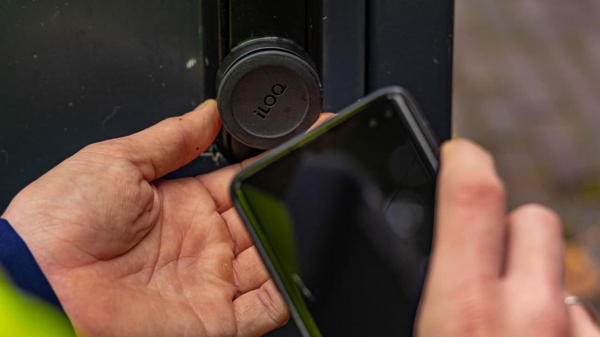 Eine Person hält ein Smartphone in die Nähe eines schwarzen iLOQ-Digitalschlosses und entriegelt möglicherweise das System mit dem Telefon, während die andere Hand das Schloss festhält.