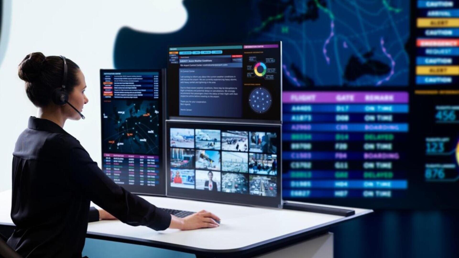 A woman wearing a headset sits at a desk surrounded by multiple computer monitors displaying maps, security camera feeds, data charts, and flight information in a control room setting.