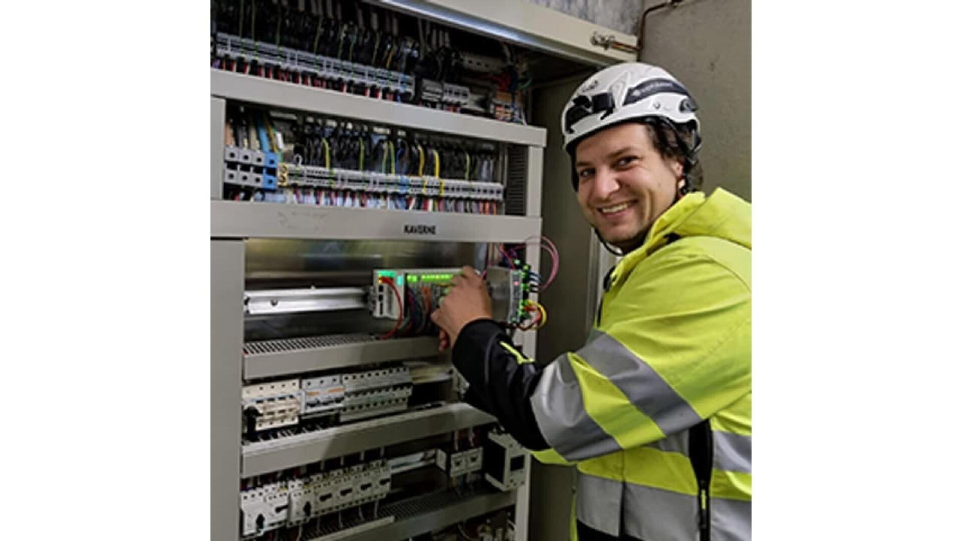 A smiling worker in a reflective jacket and helmet stands in front of an open electrical control panel, adjusting wires and components inside the cabinet.
