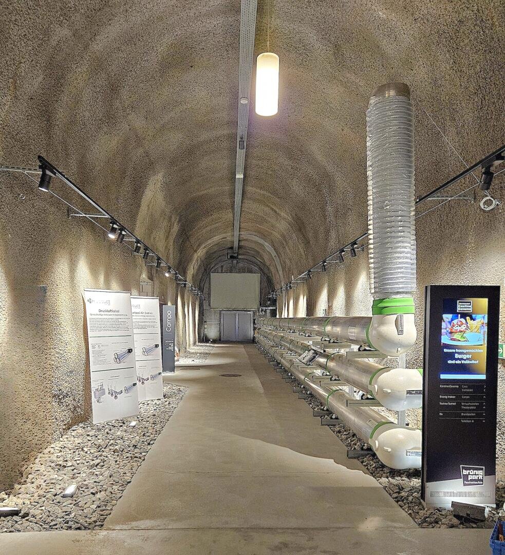 Ein tunnelartiger Raum mit Betonwänden, Informationstafeln auf der linken Seite, einem hohen metallenen Belüftungsrohr auf der rechten Seite und großen horizontalen Rohren mit Messgeräten auf der rechten Seite; Oberlichter beleuchten den Raum.