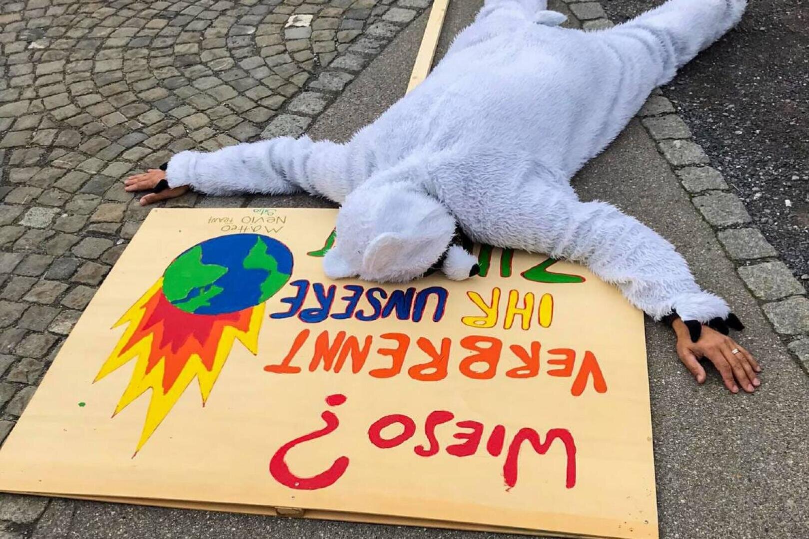 Eine Person in einem pelzigen Tierkostüm liegt mit dem Gesicht nach unten auf dem Bürgersteig, die Arme ausgestreckt, neben einem großen Schild, das eine brennende Erde und einen deutschen Text zeigt, möglicherweise als Protest gegen den Klimawandel. Das Schild steht für den Betrachter auf dem Kopf.