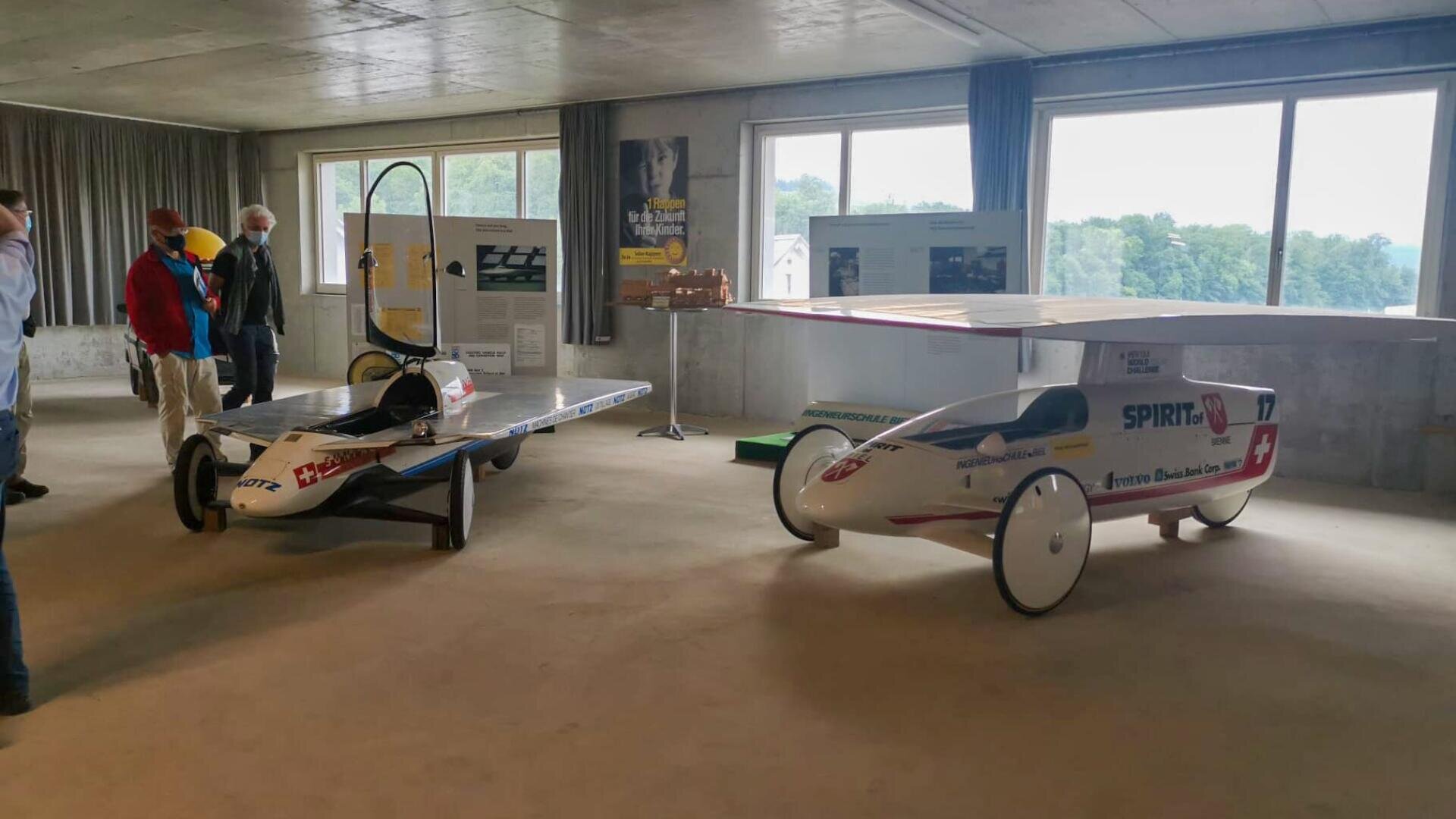 Zwei solarbetriebene Rennwagen sind in einer Halle ausgestellt, hinter denen Informationstafeln angebracht sind. Vier Personen stehen in der Nähe und beobachten die Fahrzeuge. Große Fenster lassen das Tageslicht herein und zeigen die Bäume draußen.